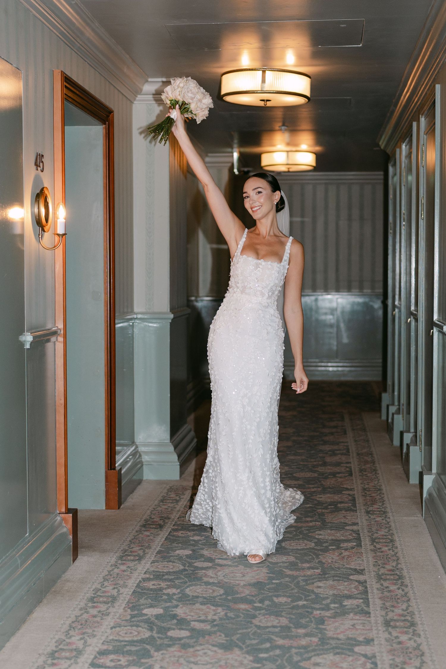 A bride in a white lace gown poses playfully in an elegant hotel hallway with patterned carpet.