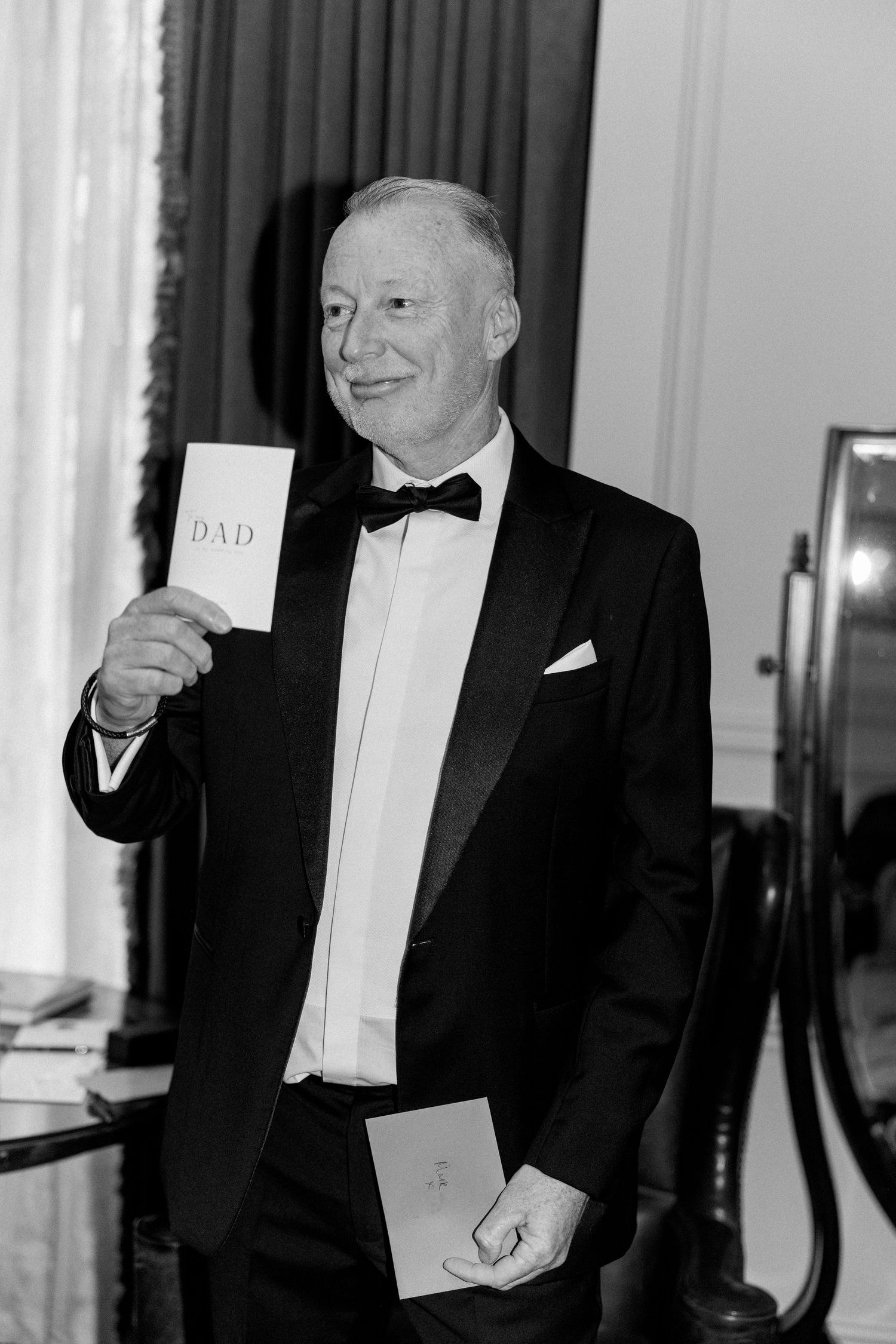 A formal event attendee in a tuxedo displays cards while standing indoors.