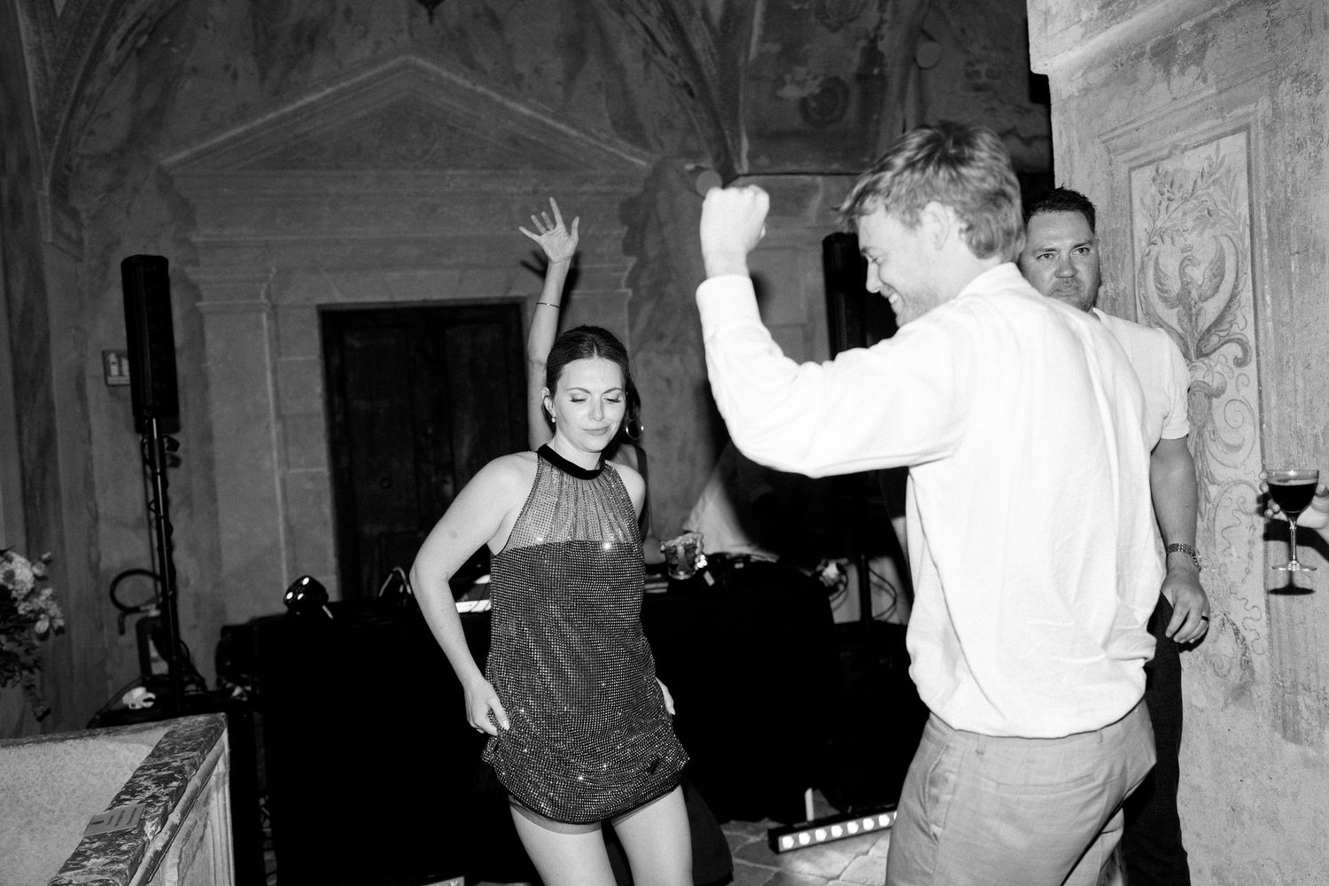 Black and white photo of partygoers dancing on a stone floor in an ornate interior space.