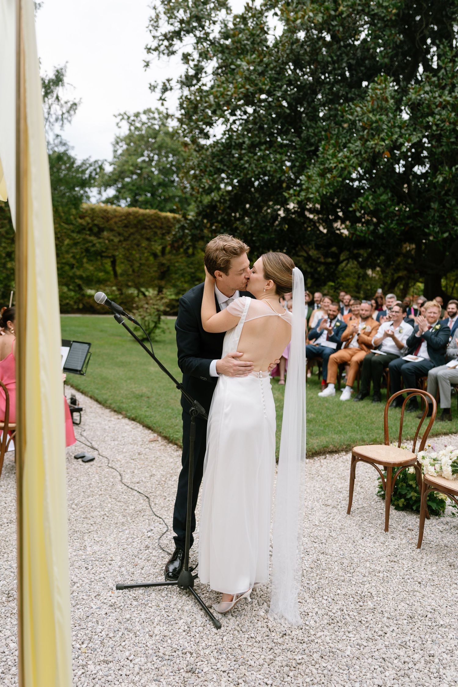 A wedding couple shares an intimate kiss during their outdoor ceremony while guests look on from garden seating.