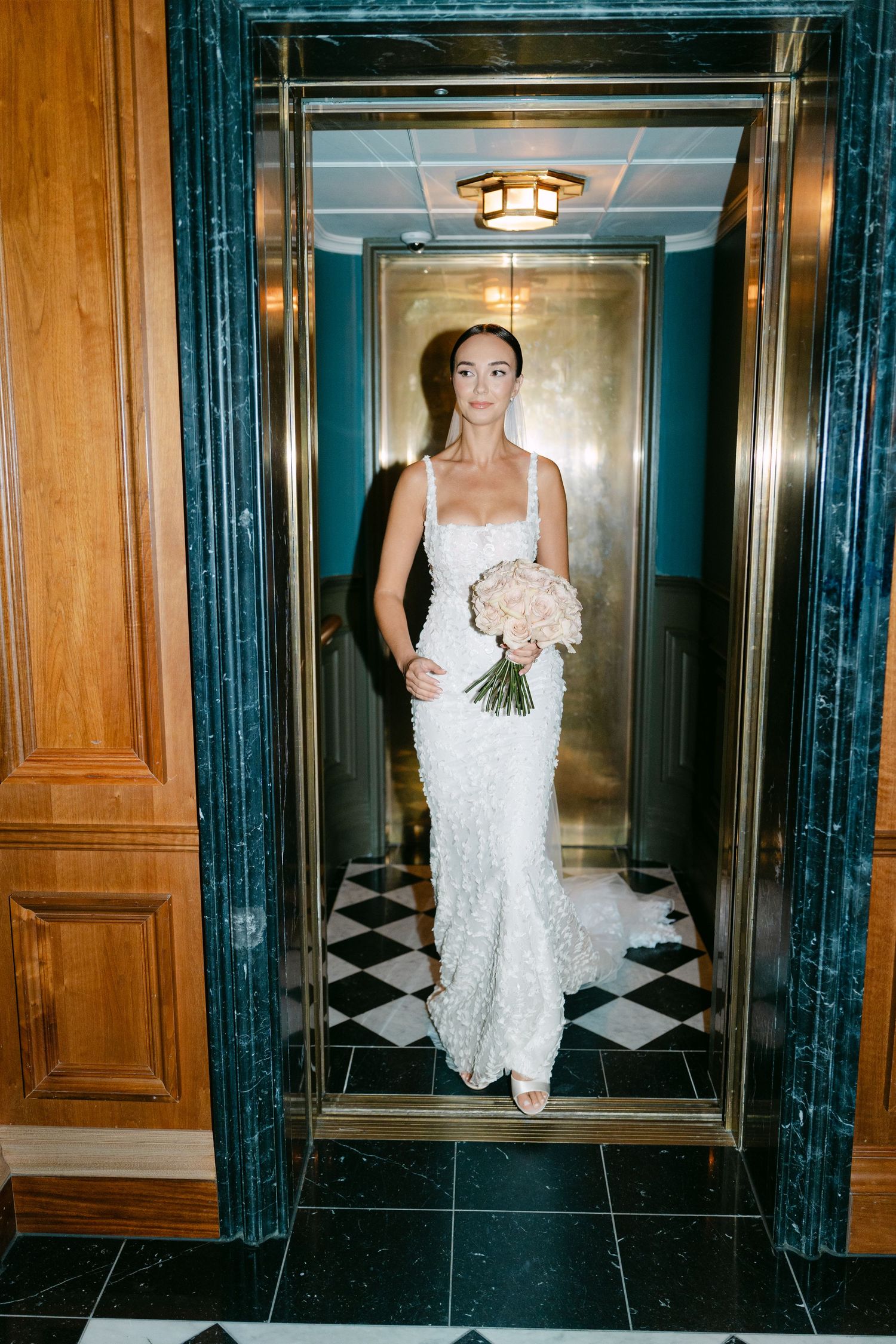 Bride in fitted lace gown stands in elegant hallway with marble details and wooden panels.