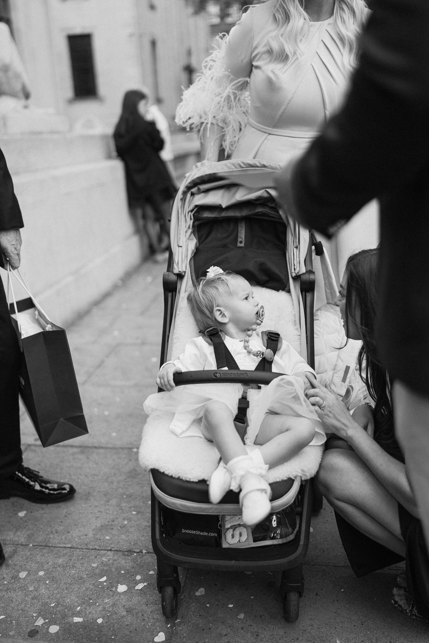 Child in stroller at wedding celebration surrounded by formal attire and umbrellas.