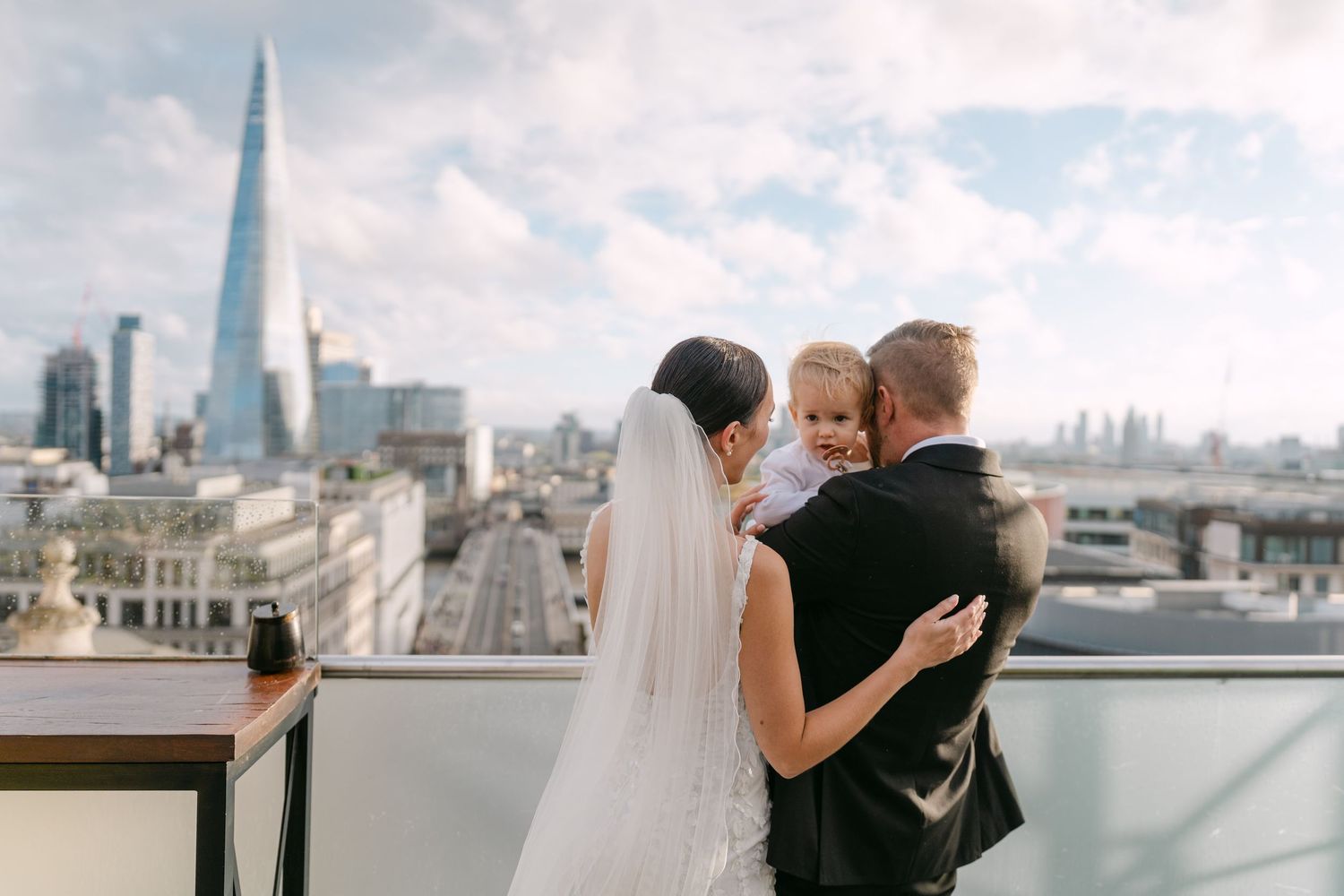 A wedding couple embraces on a rooftop terrace with London's Shard building visible in the skyline.