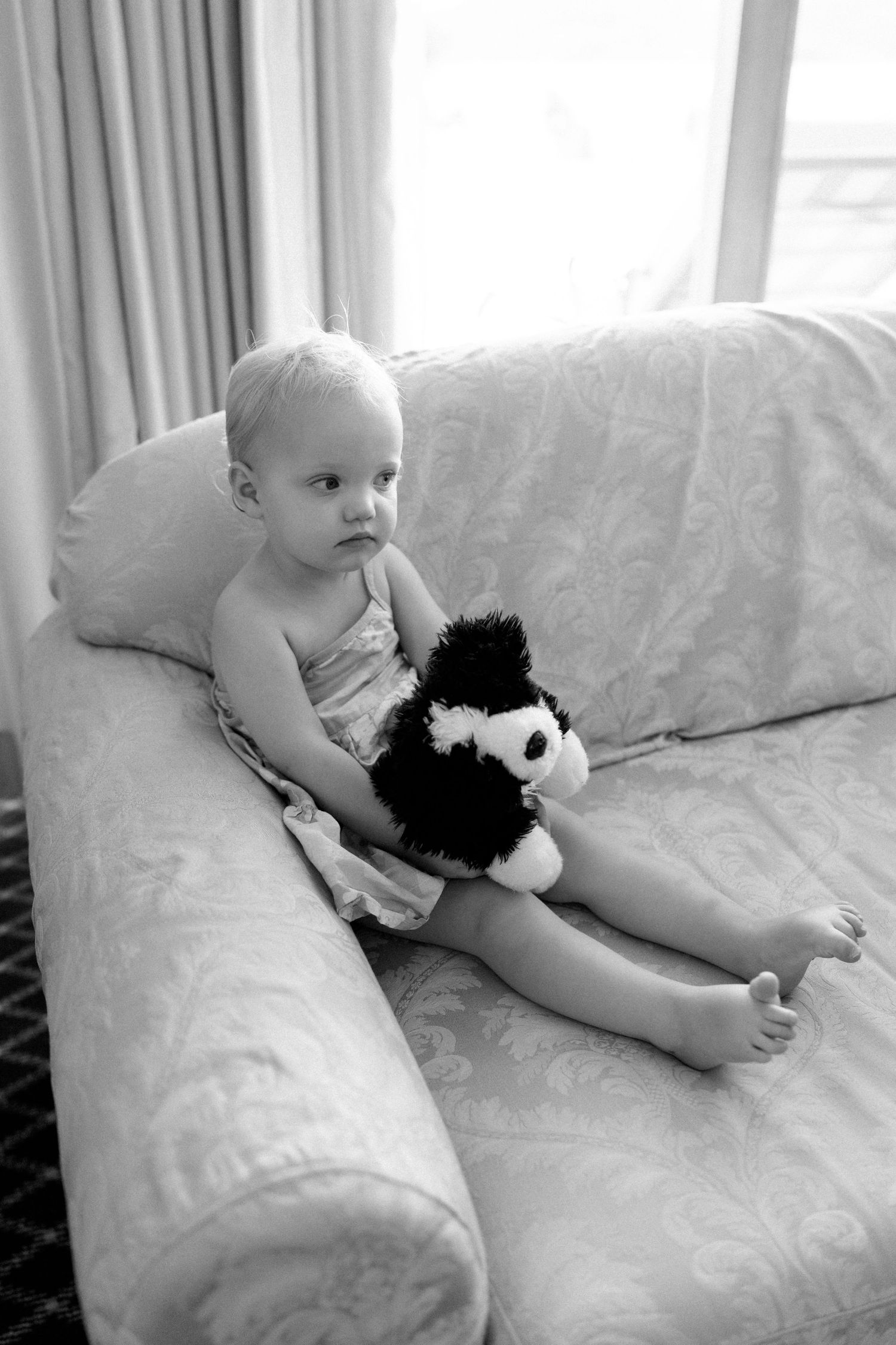 Baby in white diaper lying on white couch with black stuffed animal.