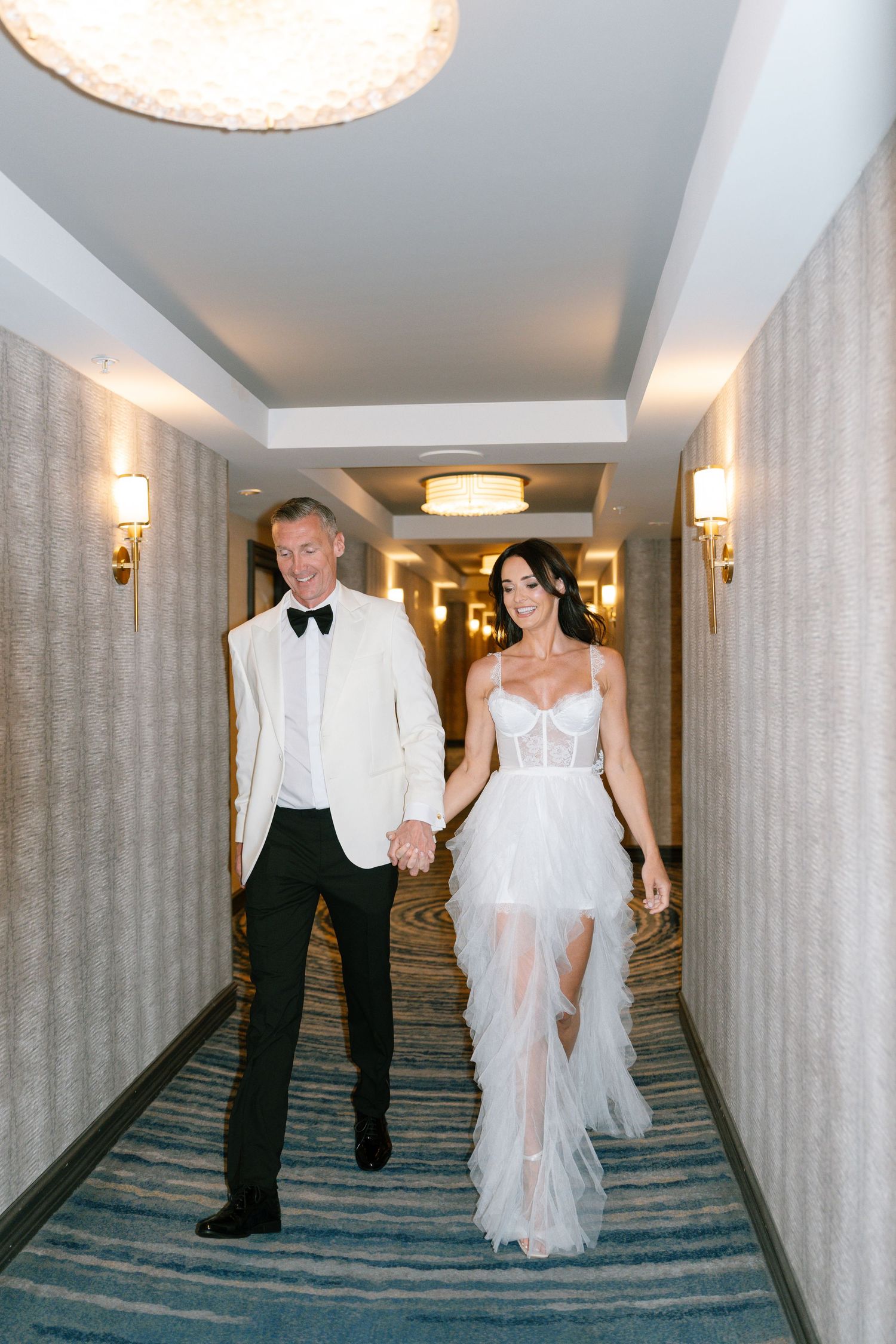 Couple walking hand in hand down elegant hotel hallway in wedding attire.