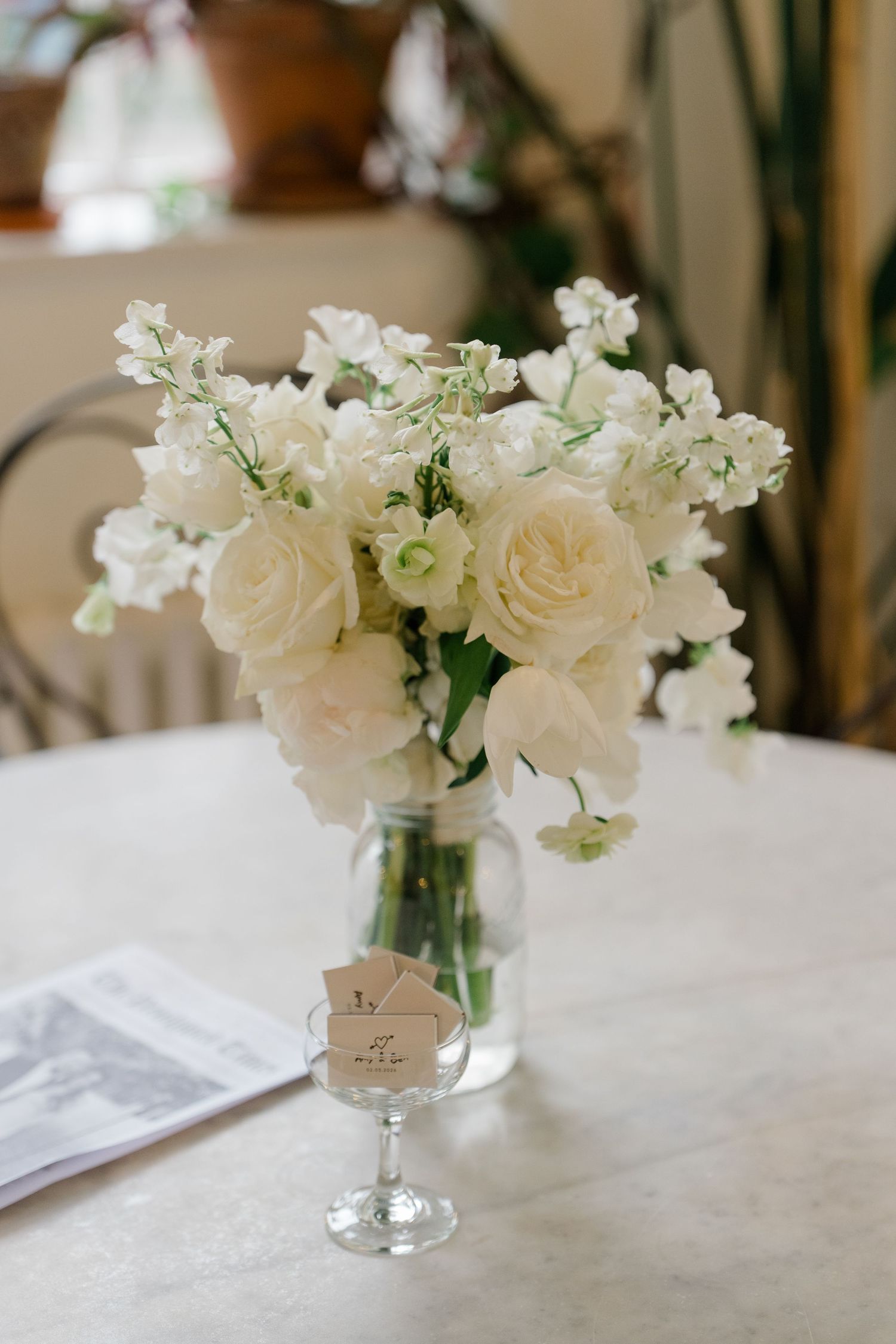 White roses and sweet peas in a glass vase on a marble table with a wedding program and place card.