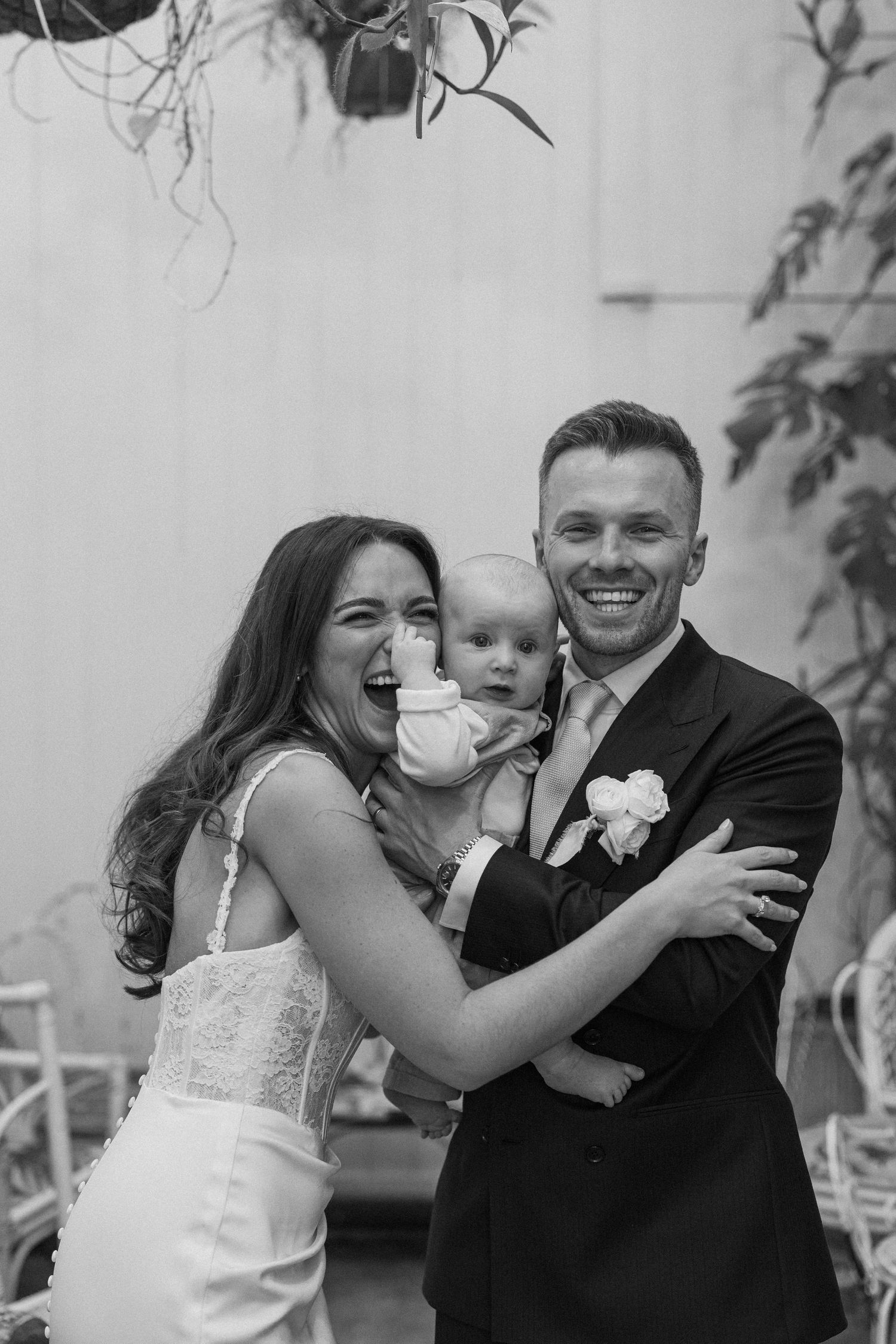 Black and white photo of smiling bride and groom holding baby at wedding ceremony.