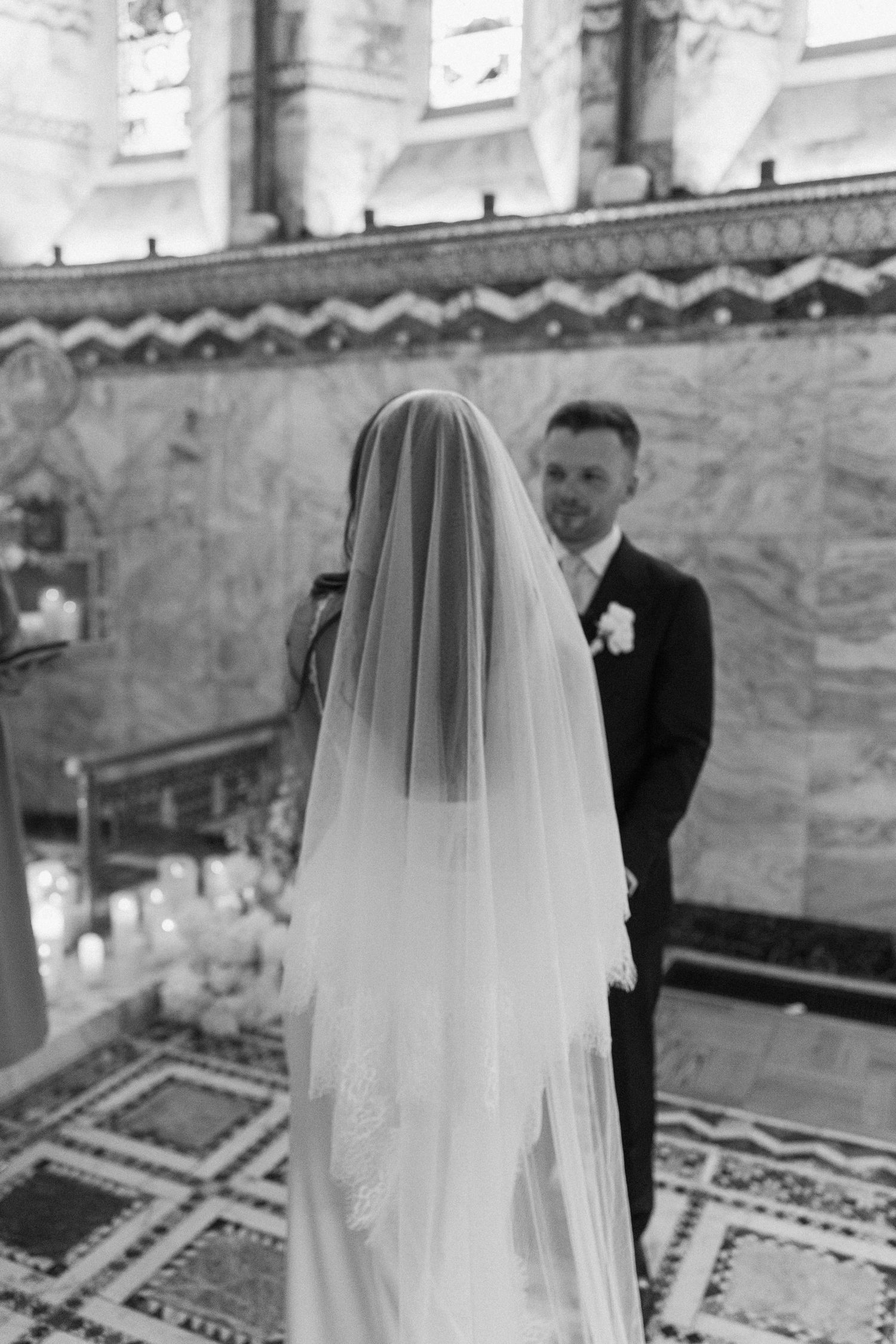 Bride in lace wedding dress and veil stands at church altar facing groom during wedding ceremony.