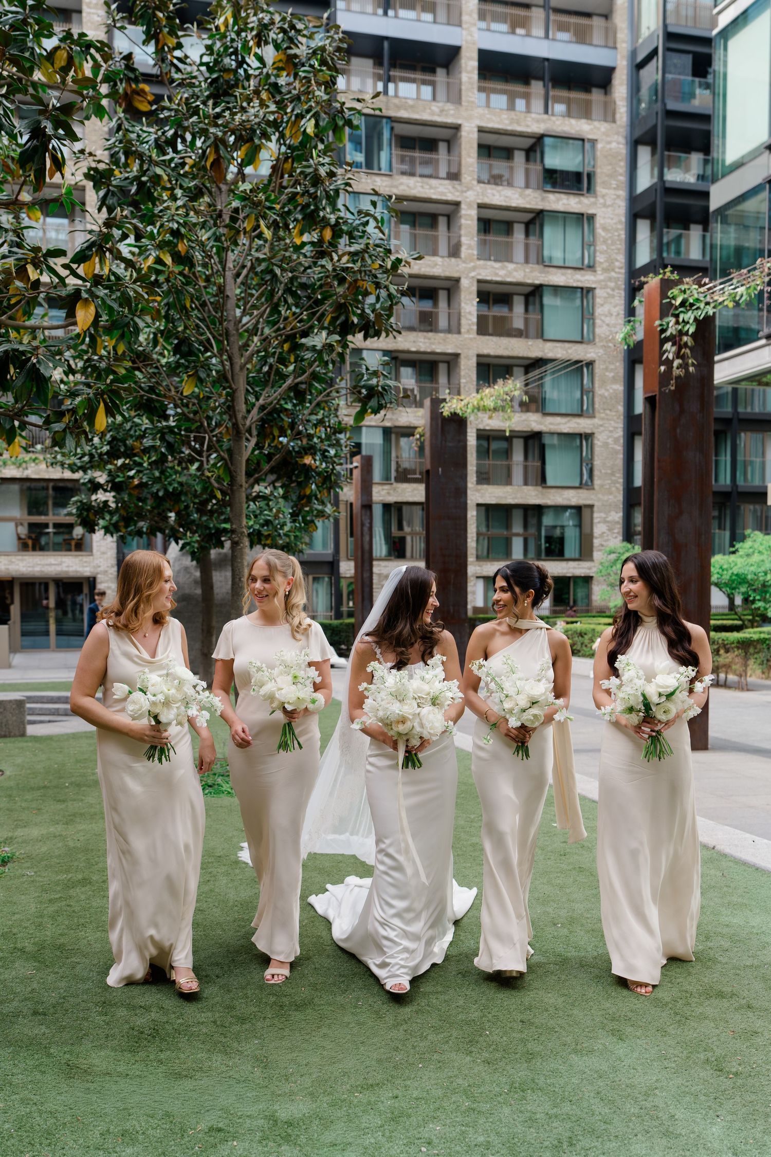 Bride in white gown walks with four bridesmaids in champagne dresses holding white bouquets outdoors.