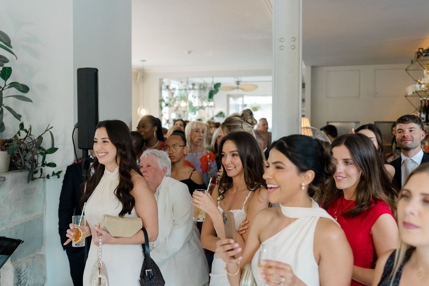 Wedding guests in white and red dresses mingle and laugh during cocktail hour in bright modern venue.
