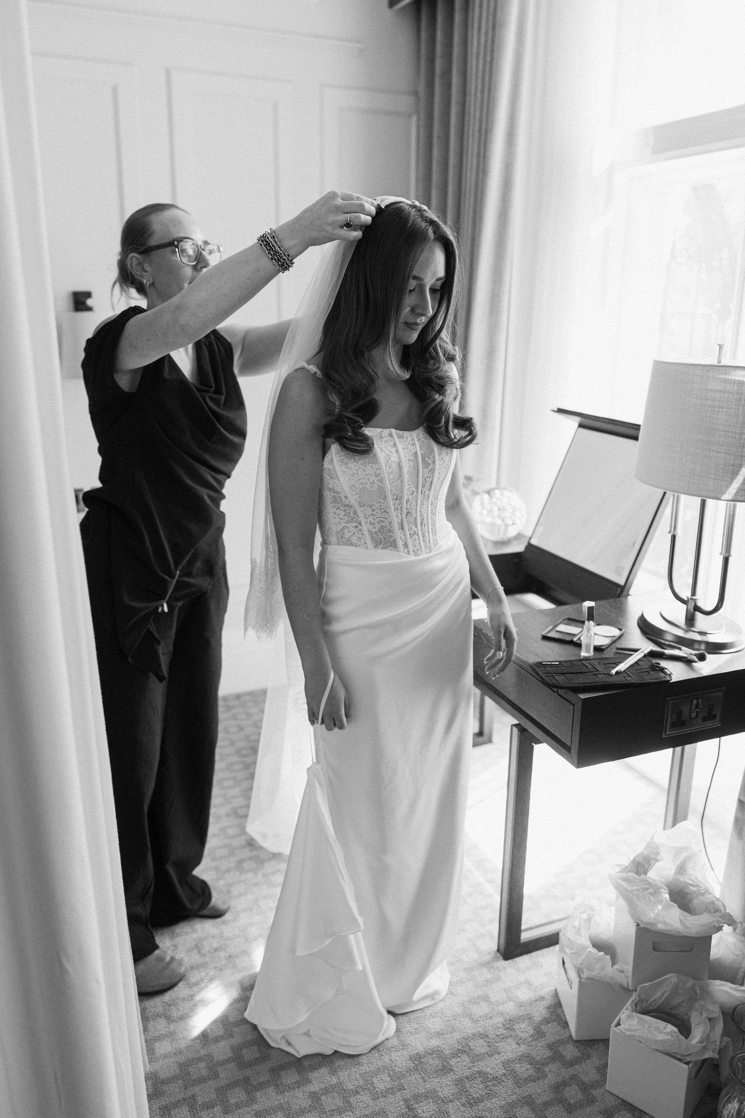 Black and white photo of hairdresser placing veil on smiling bride in lace wedding dress.