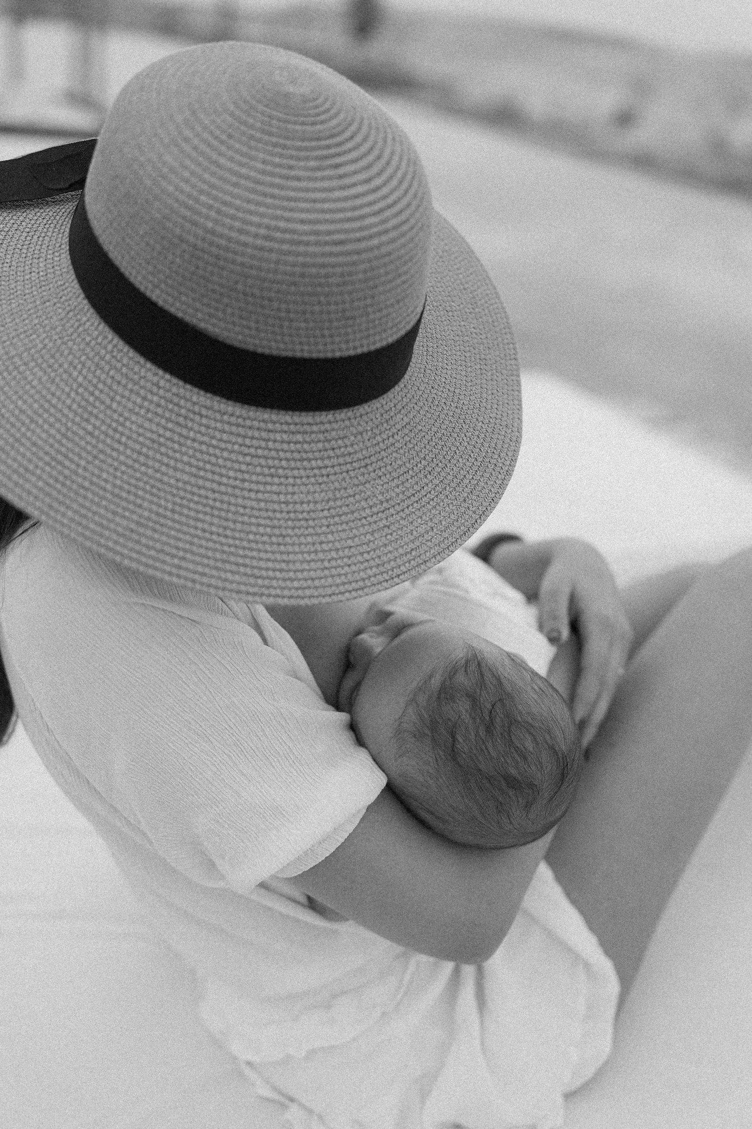 Black and white photo of a mother in a wide-brim sun hat breastfeeding her newborn baby outdoors.