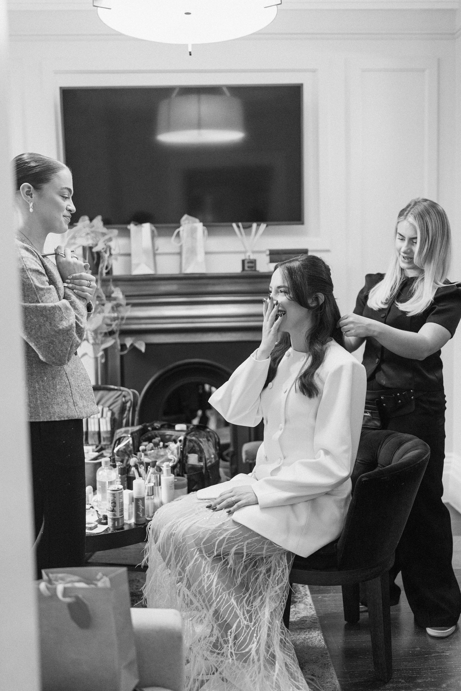 Bride getting hair styled in black and white photo with makeup artist and friend in elegant room.