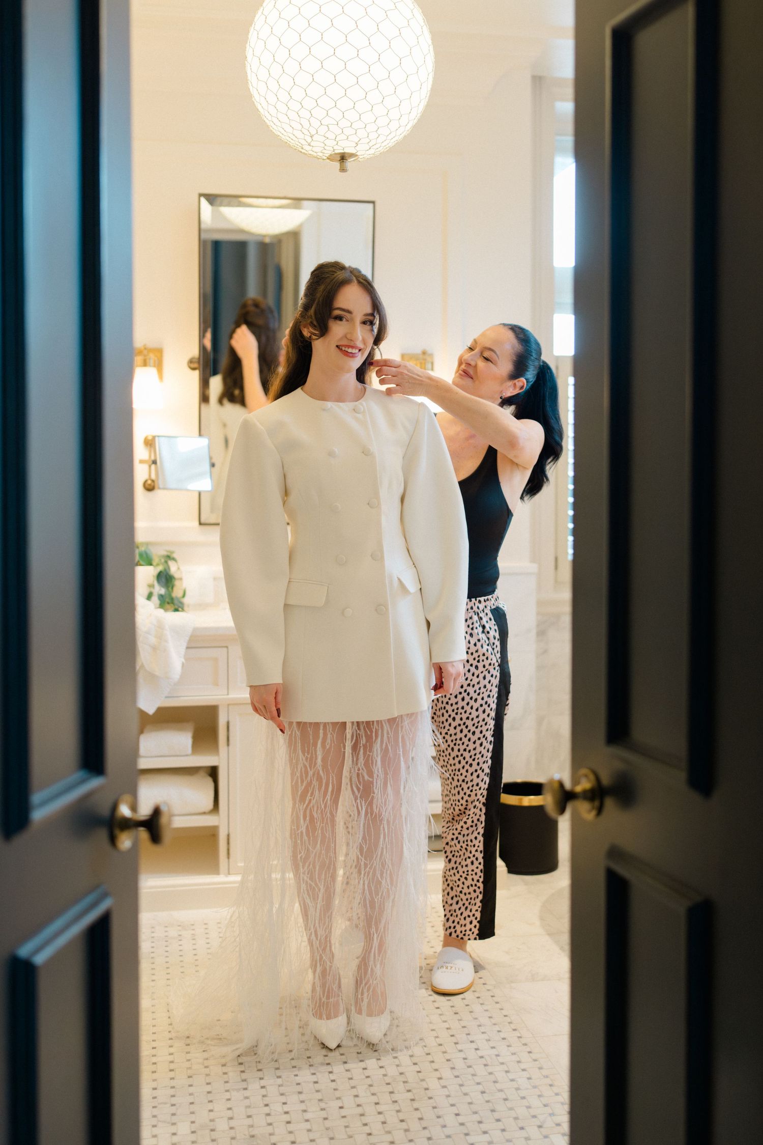 Bride in white blazer dress with feather skirt being helped by bridesmaid in elegant hotel suite.