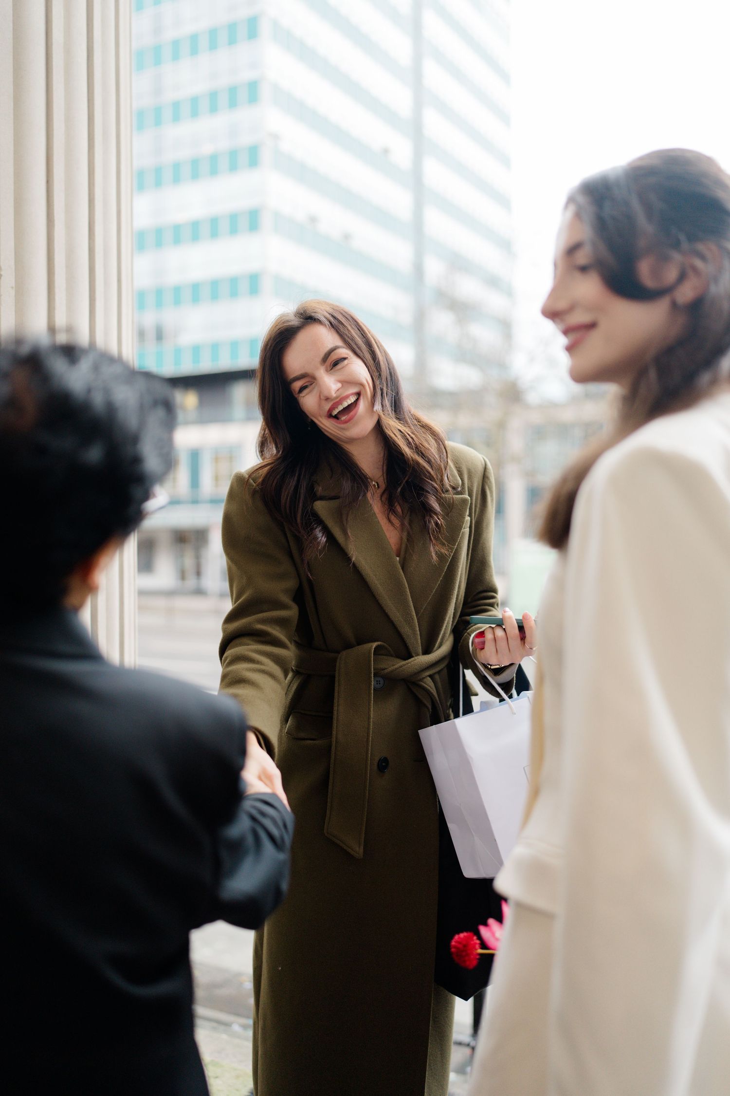 Smiling woman in olive coat laughs while holding shopping bags outside modern city building.