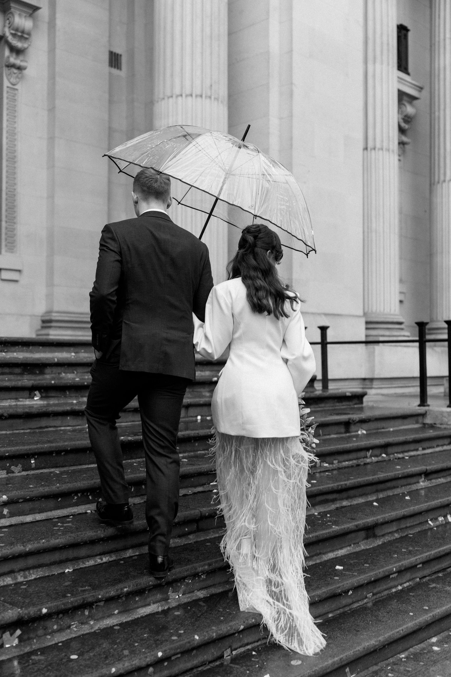 Bride and groom walking up stone steps sharing clear umbrella in black and white wedding photo.