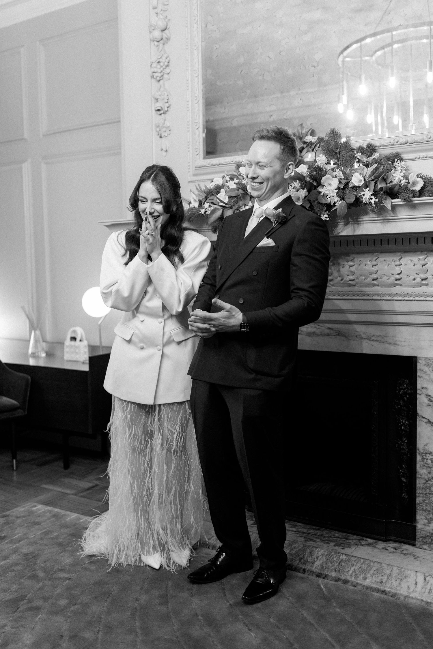 Black and white photo of laughing bride and groom standing before ornate fireplace at wedding.