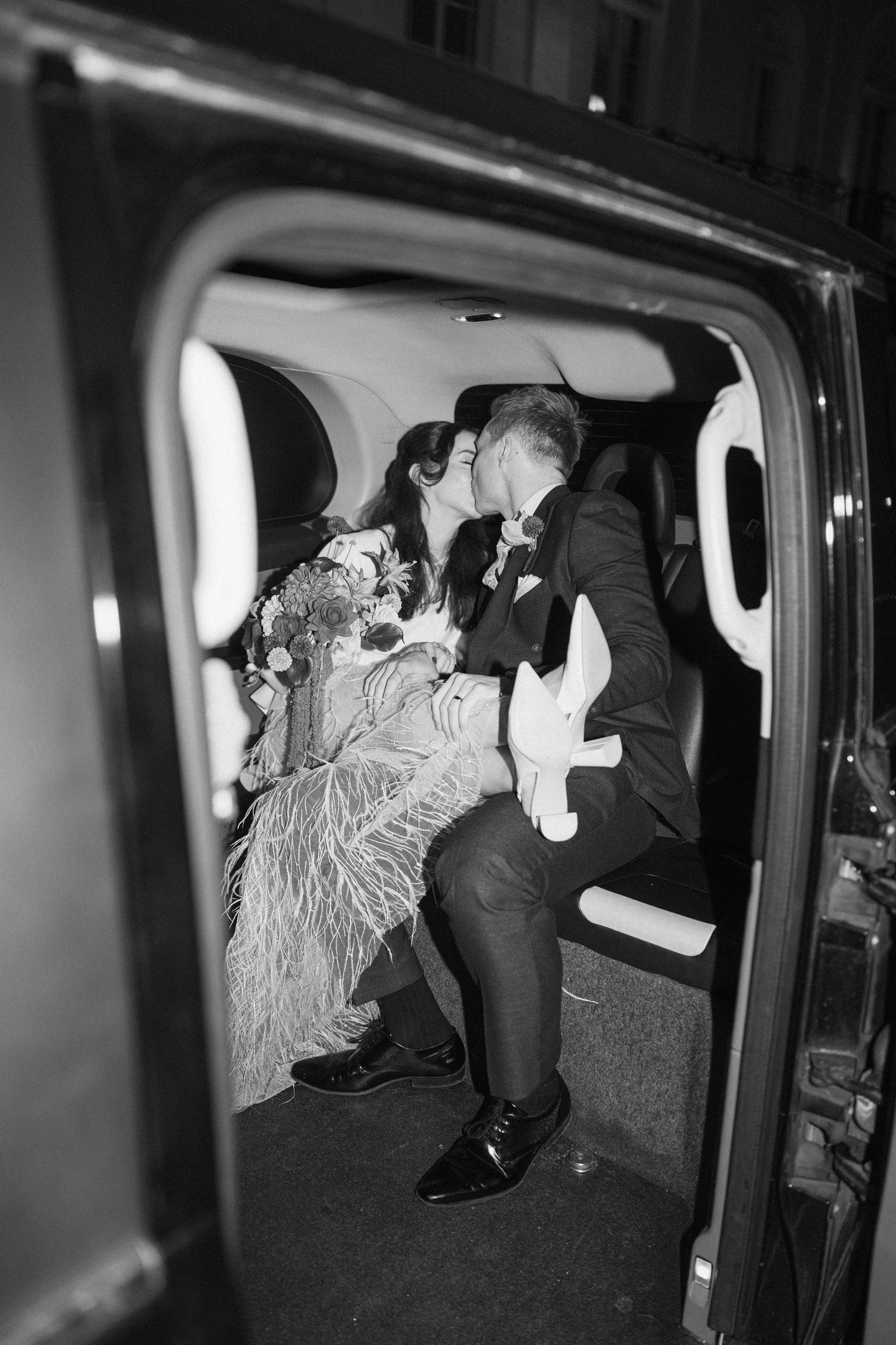 Bride and groom kissing inside black cab on wedding night, black and white photo.