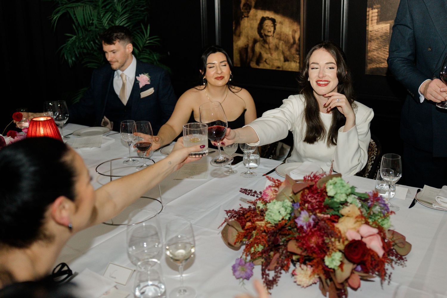 Bride in white dress toasting with guests at a wedding reception table adorned with colorful floral centerpieces.
