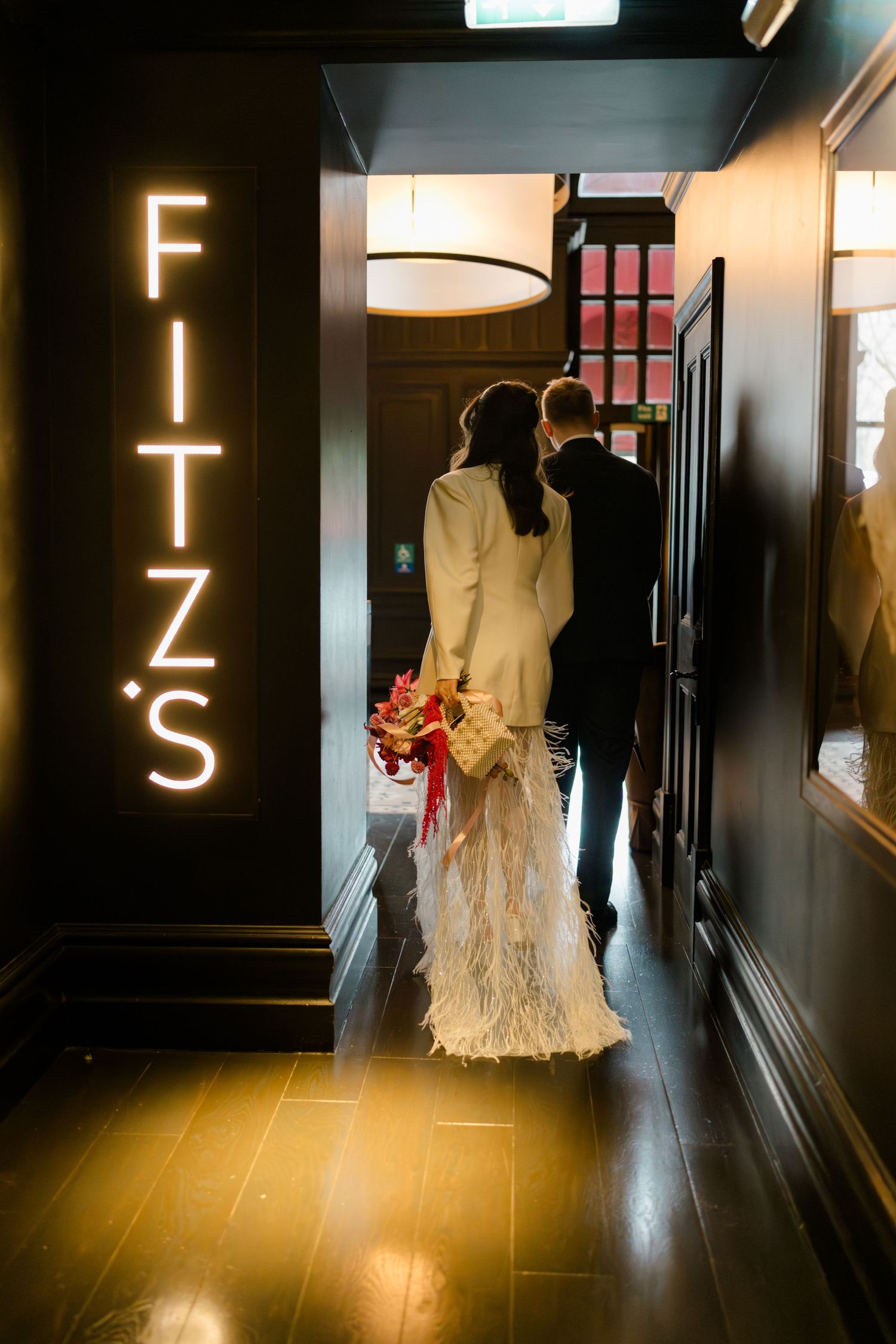 Newlyweds walking hand in hand past illuminated Fitz's neon sign at wedding venue.