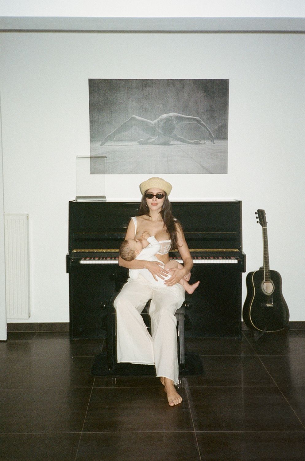 Woman in white outfit and beret breastfeeding baby in front of black upright piano with guitar nearby.