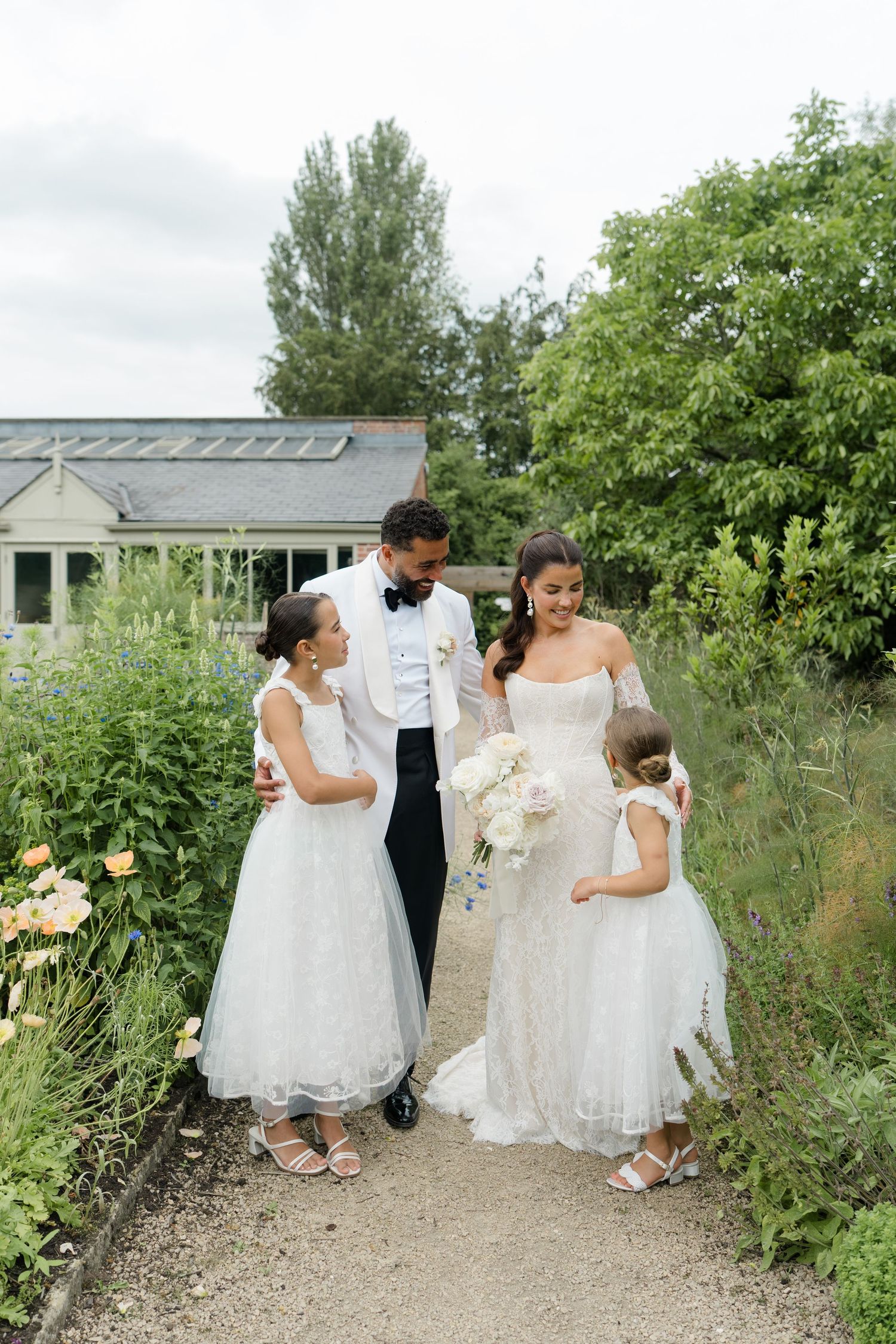 Bride and groom pose with two flower girls in white dresses along a garden path at outdoor wedding.