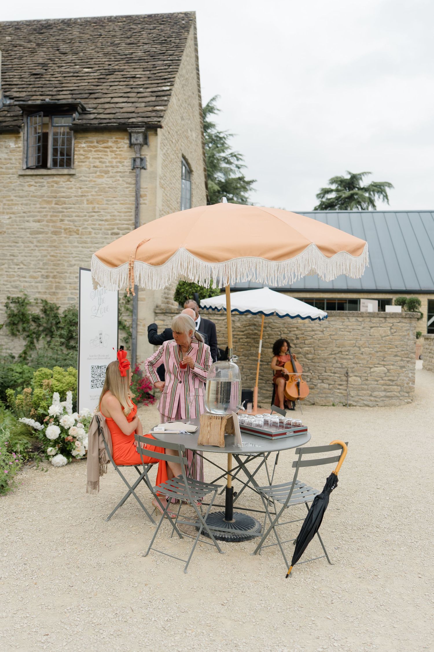 Guests gather at outdoor welcome table under peach fringed umbrella at stone manor wedding venue.