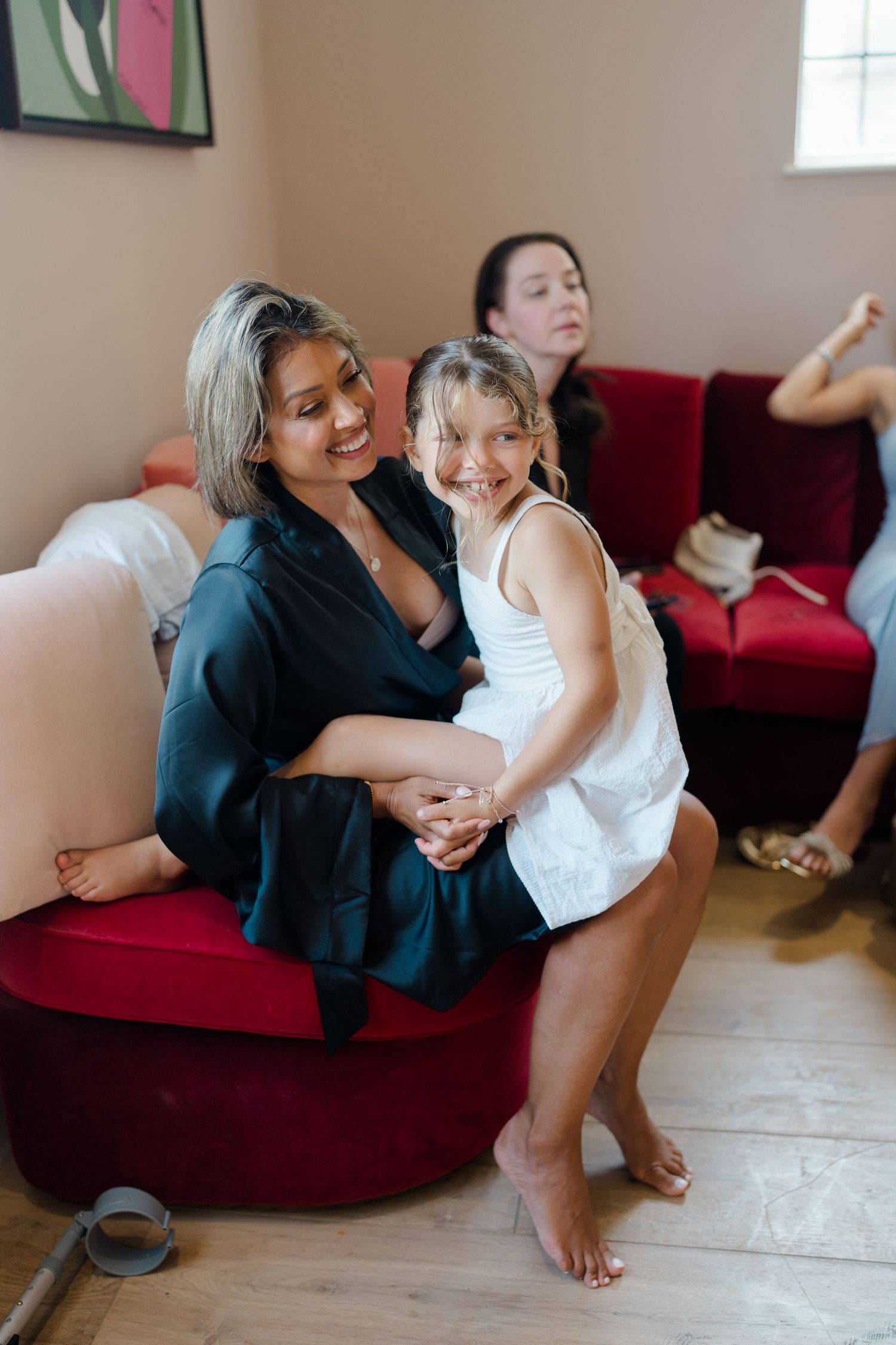 A smiling woman in a black satin robe hugs a young girl in white during wedding preparations on a red sofa.