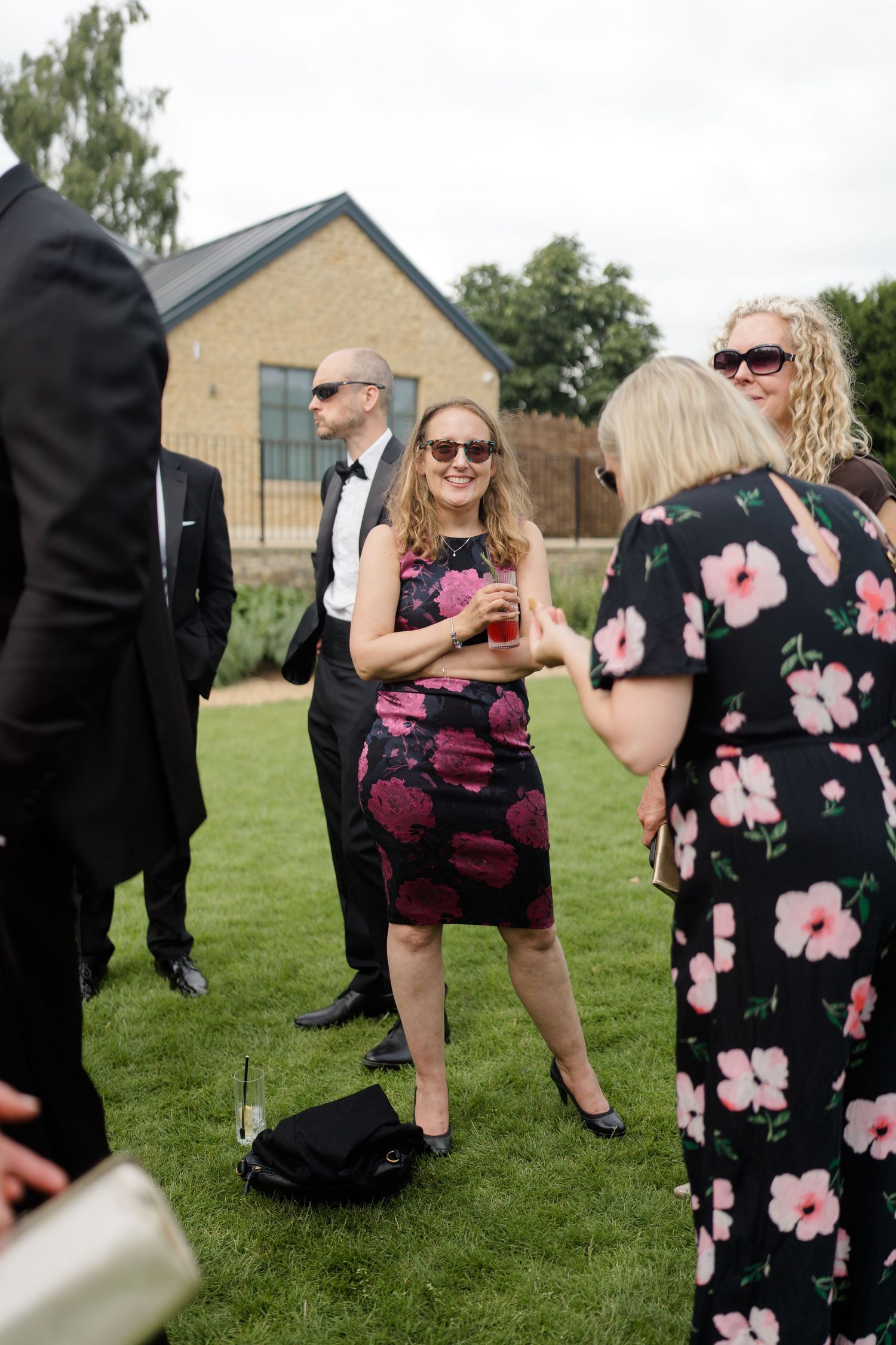 Wedding guest in black floral dress smiles while holding drink and chatting with others on lawn.