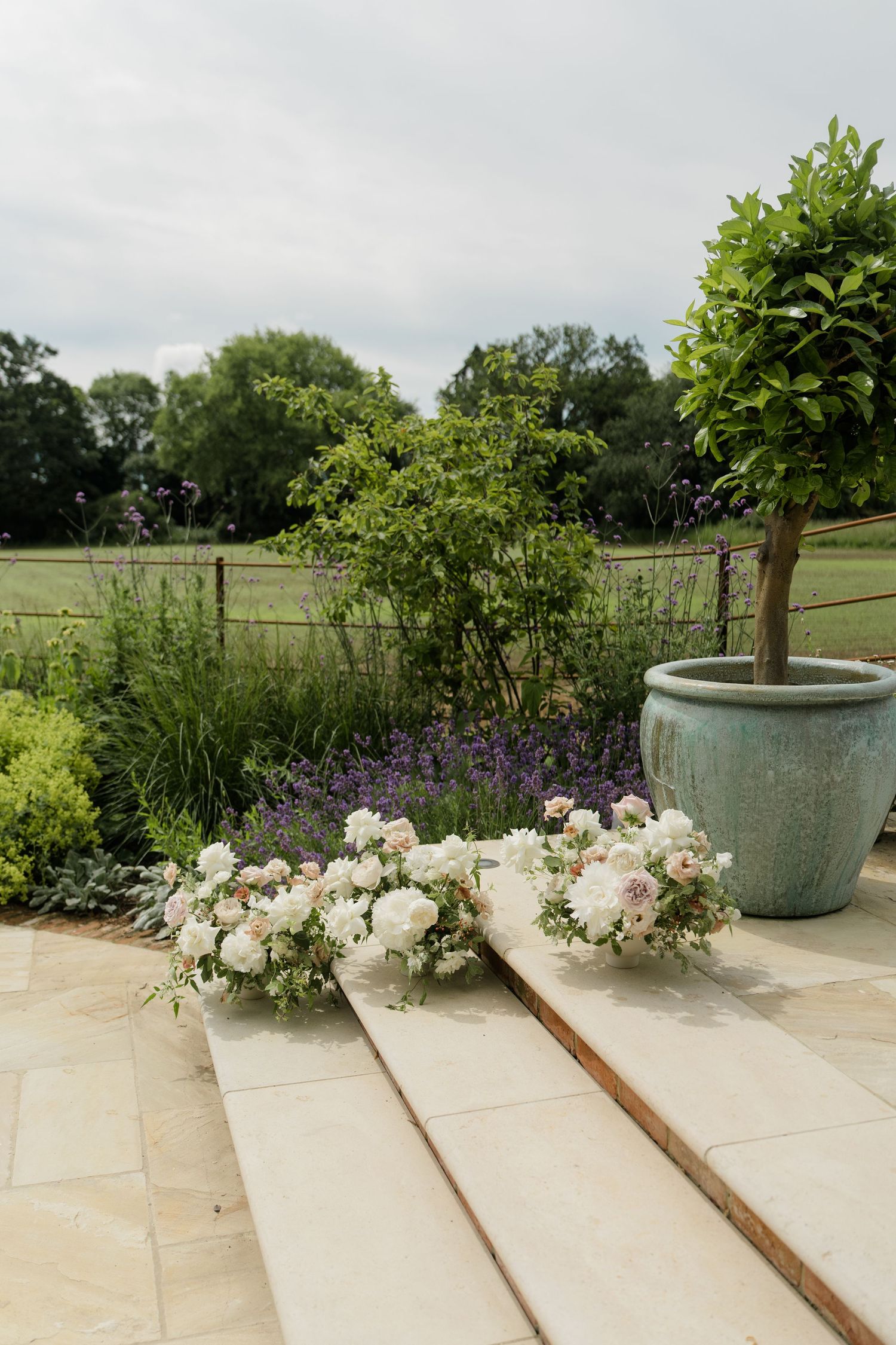 Elegant white floral wedding arrangements displayed on stone steps beside a green topiary tree.
