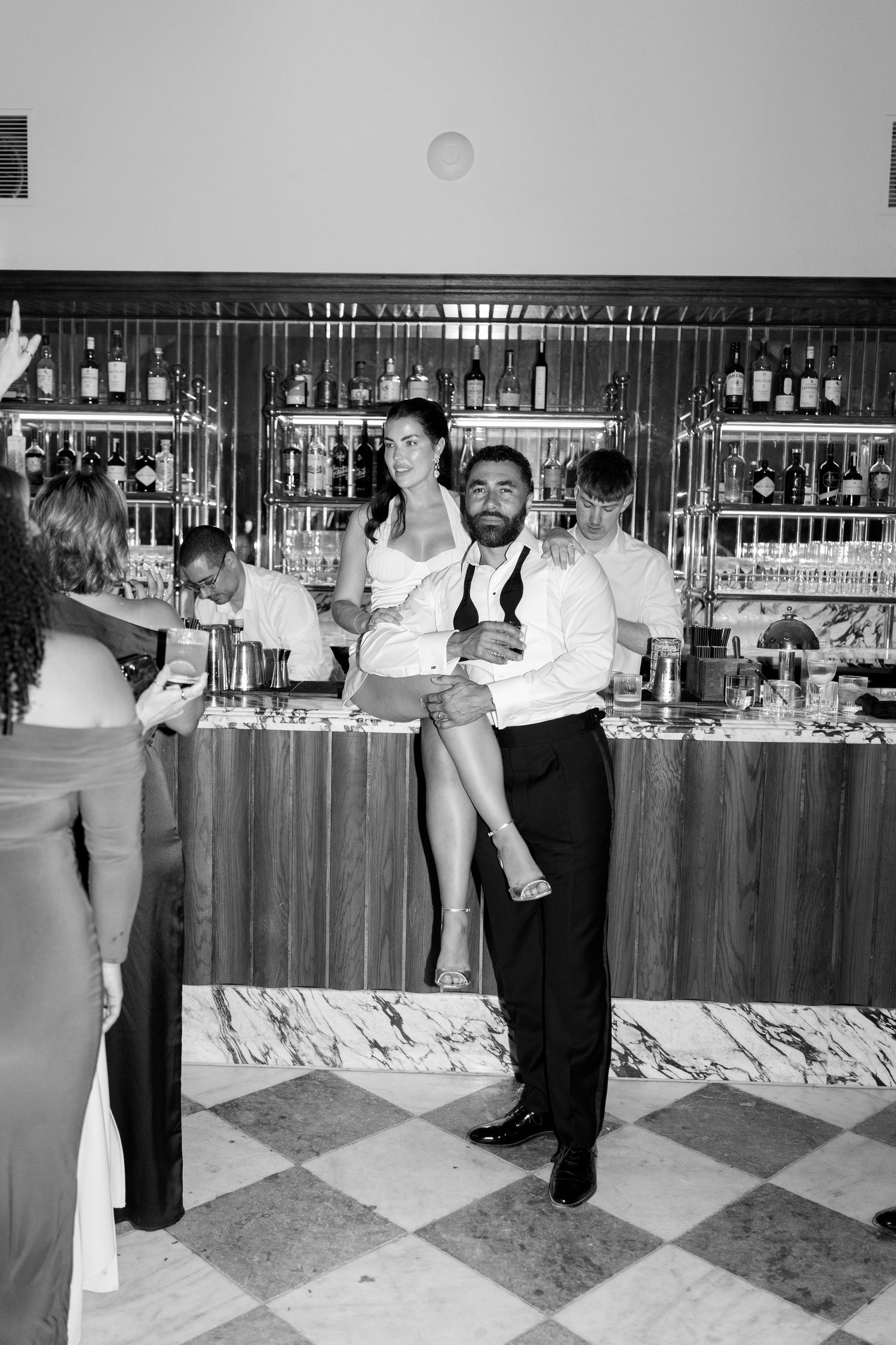 Groom carries bride at wedding bar in playful black and white reception moment.