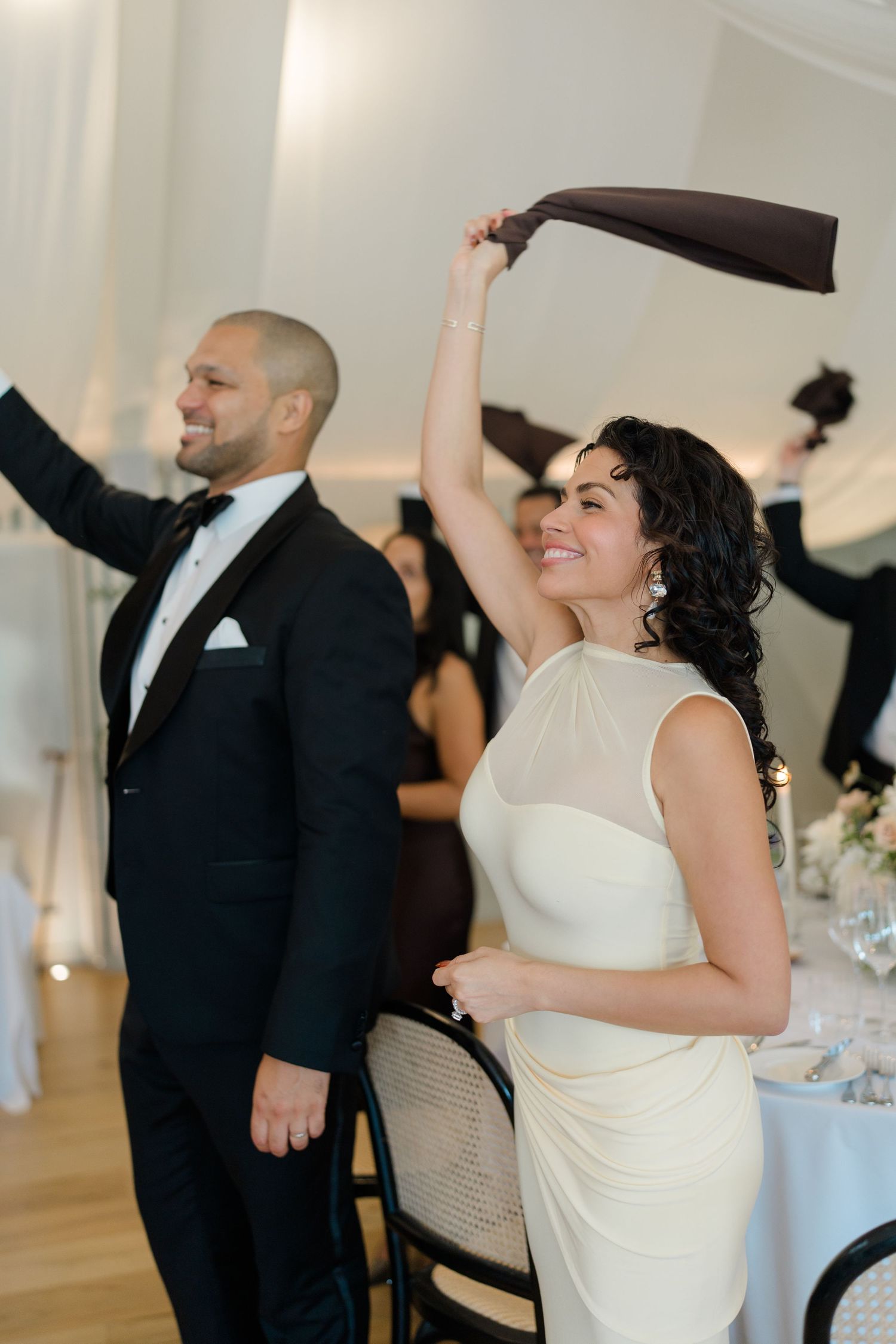 Bride in white dress and groom in tuxedo joyfully waving napkins at wedding reception dinner.