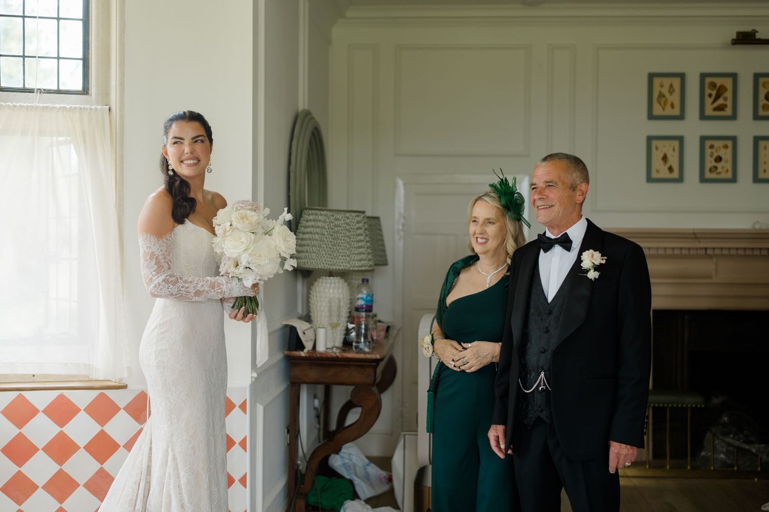 Bride in white off-shoulder gown smiling with parents in elegant room before wedding ceremony.