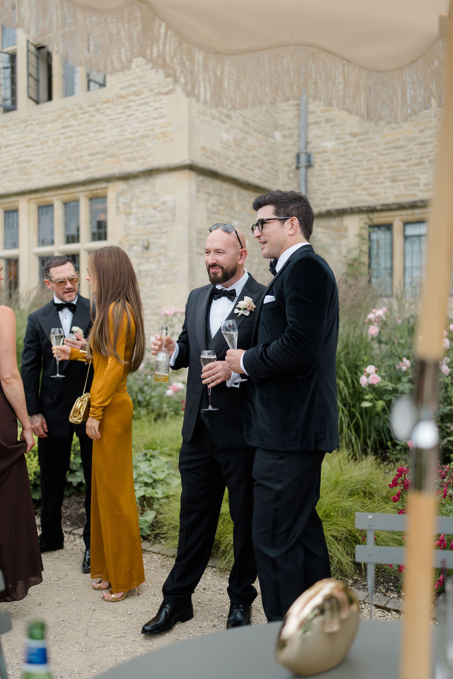 Guests in formal black tie attire socializing outdoors at a wedding reception, holding champagne glasses.