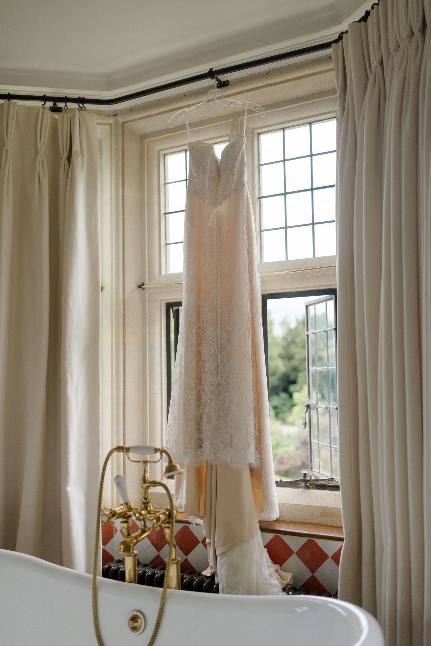 Elegant lace wedding dress hanging in a bright bathroom window with cream curtains and a freestanding bathtub.
