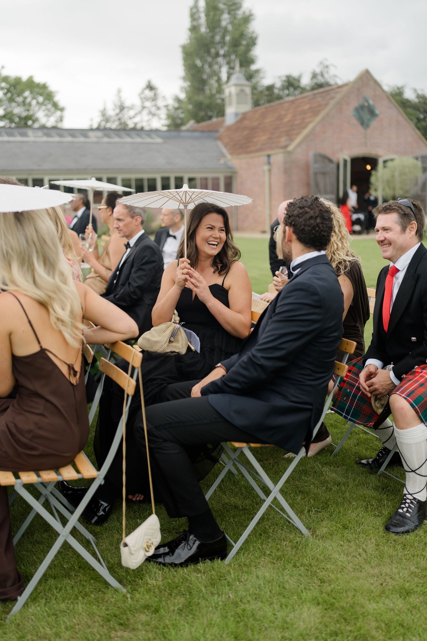 Wedding guests in formal attire laugh and chat outdoors holding parasols on garden lawn seating.