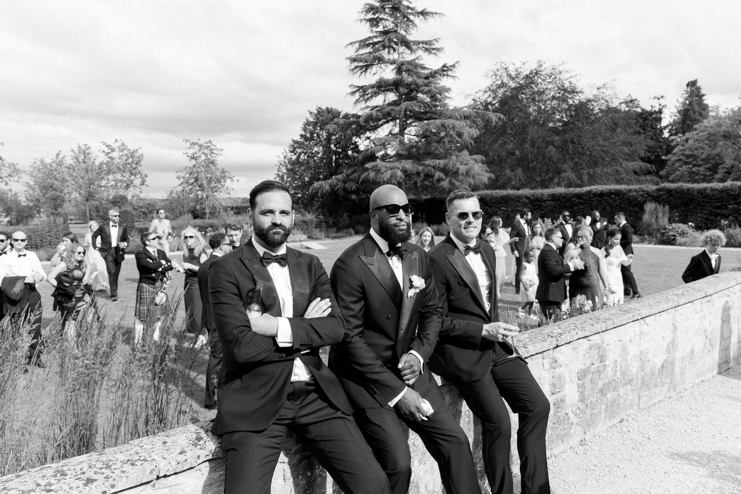 Three groomsmen in black tuxedos posing confidently at outdoor wedding reception in black and white.