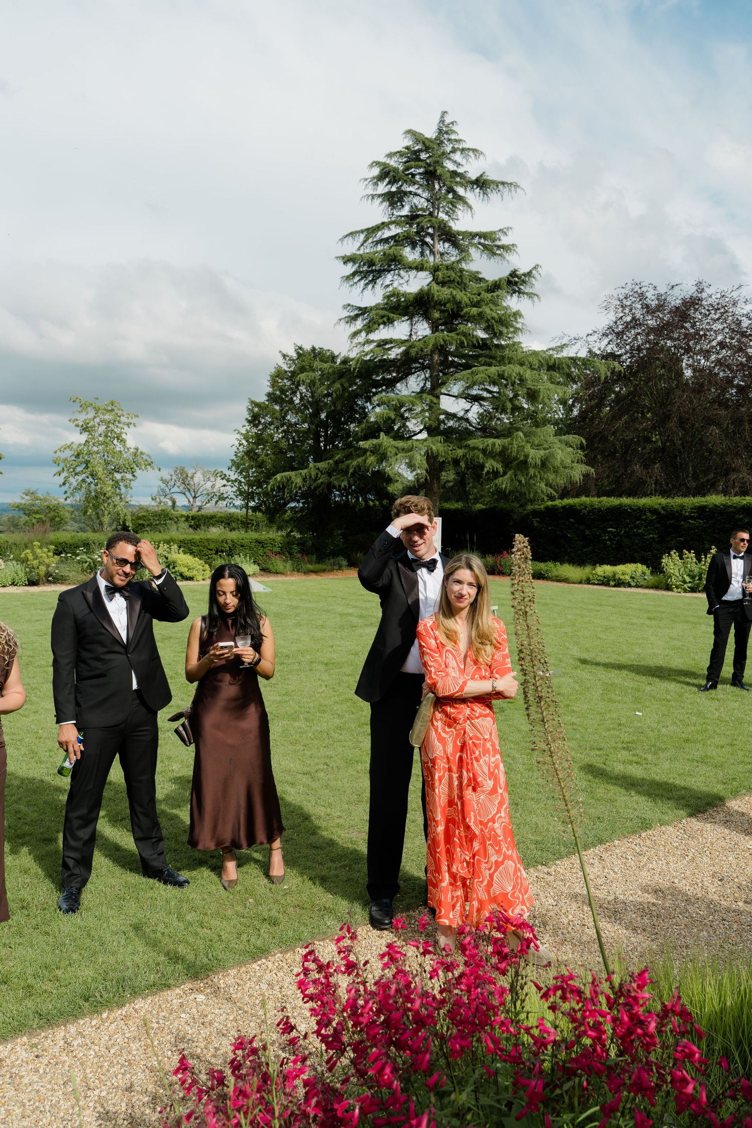 Wedding guests in formal wear enjoying outdoor reception in manicured garden of elegant estate.