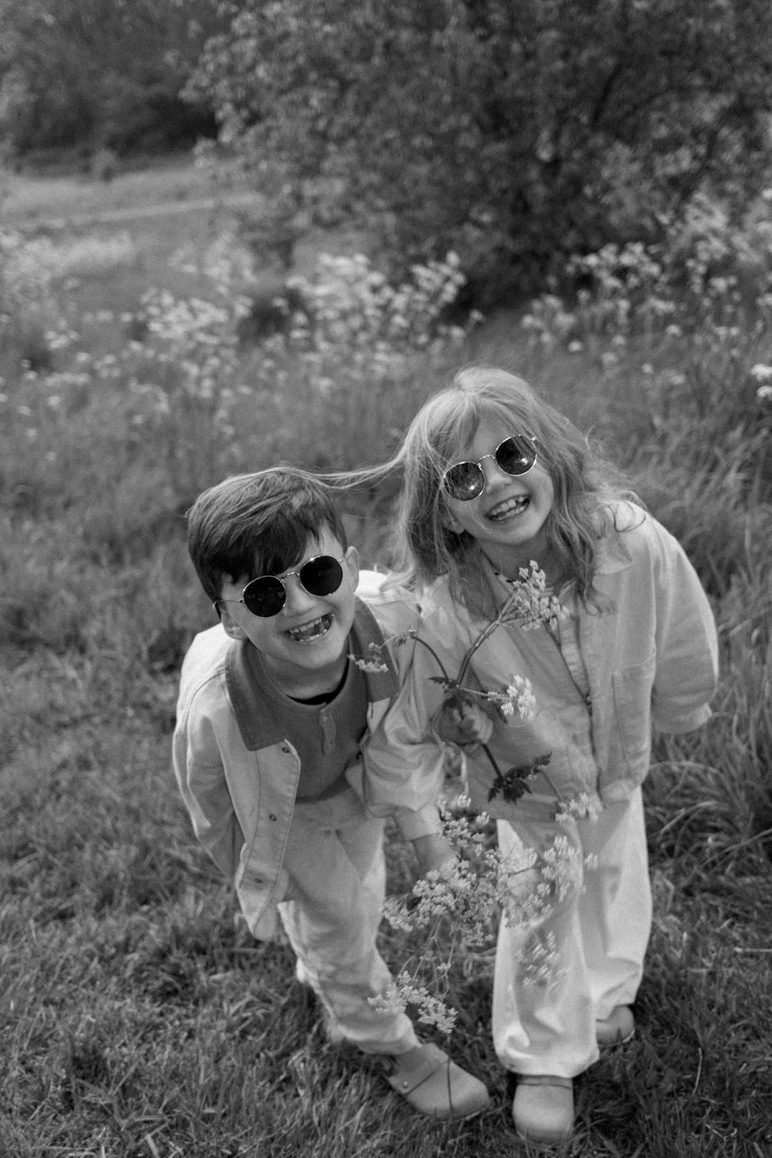 Two kids wearing sunglasses and white outfits lean together while playing in a wildflower field.