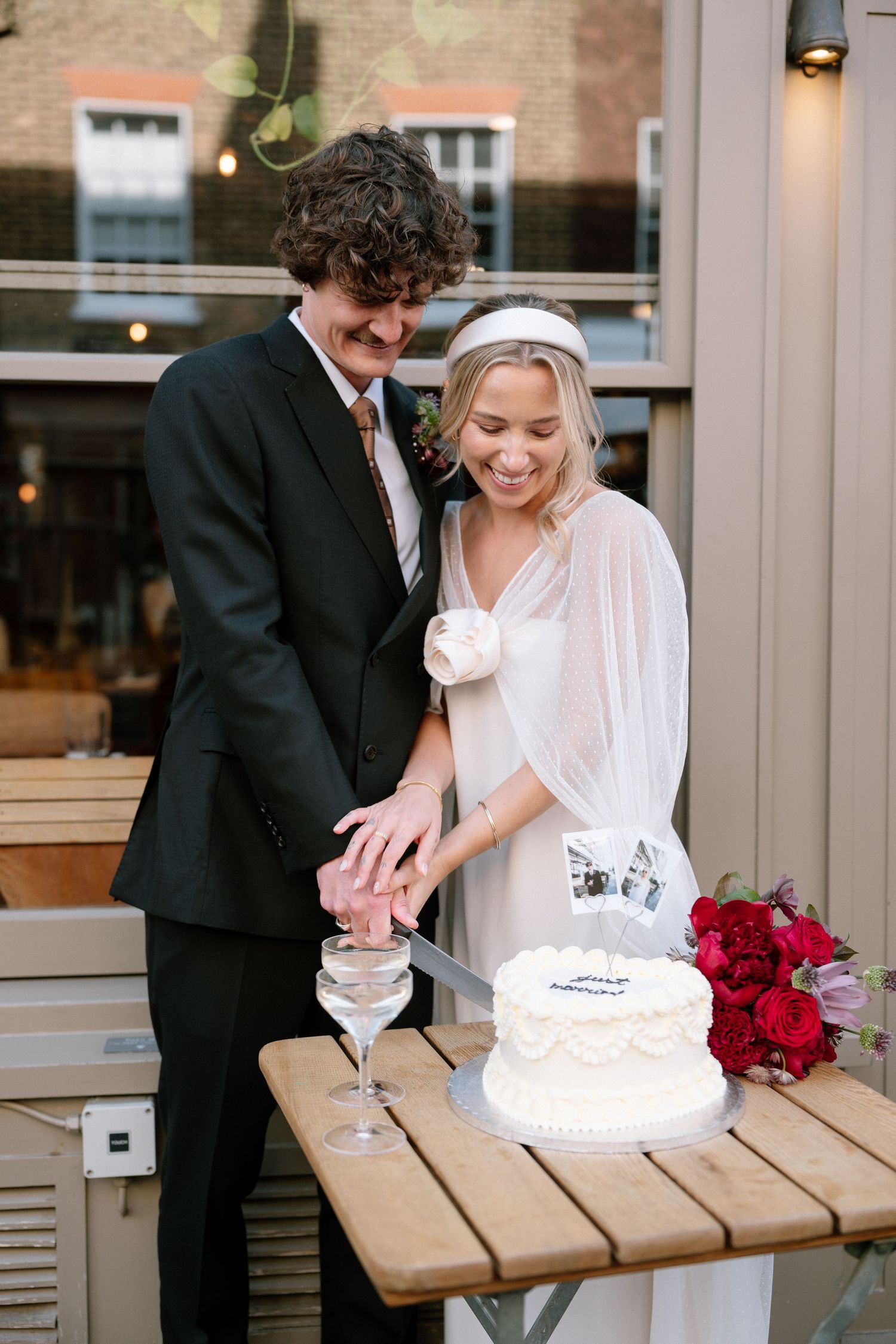 A couple cuts their wedding cake together on a wooden table with champagne and roses.