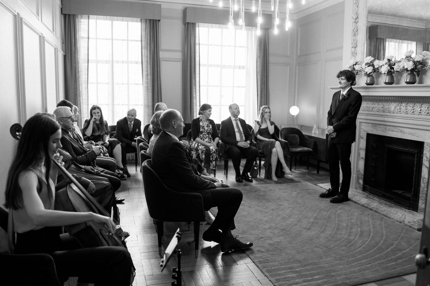Wedding guests seated in an elegant room with a fireplace during a ceremony.