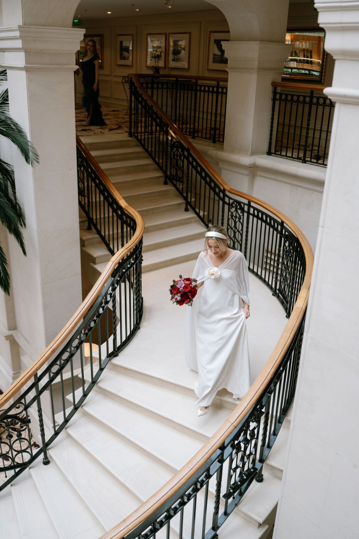 Elegant white wedding dress cascades down marble stairs with decorative black iron railings in a grand interior.