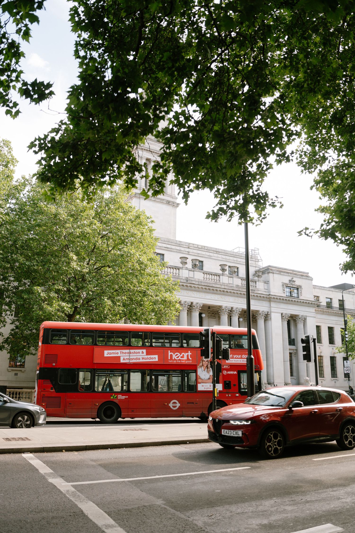Red London buses line up along a street with classic white buildings and green trees in the background.