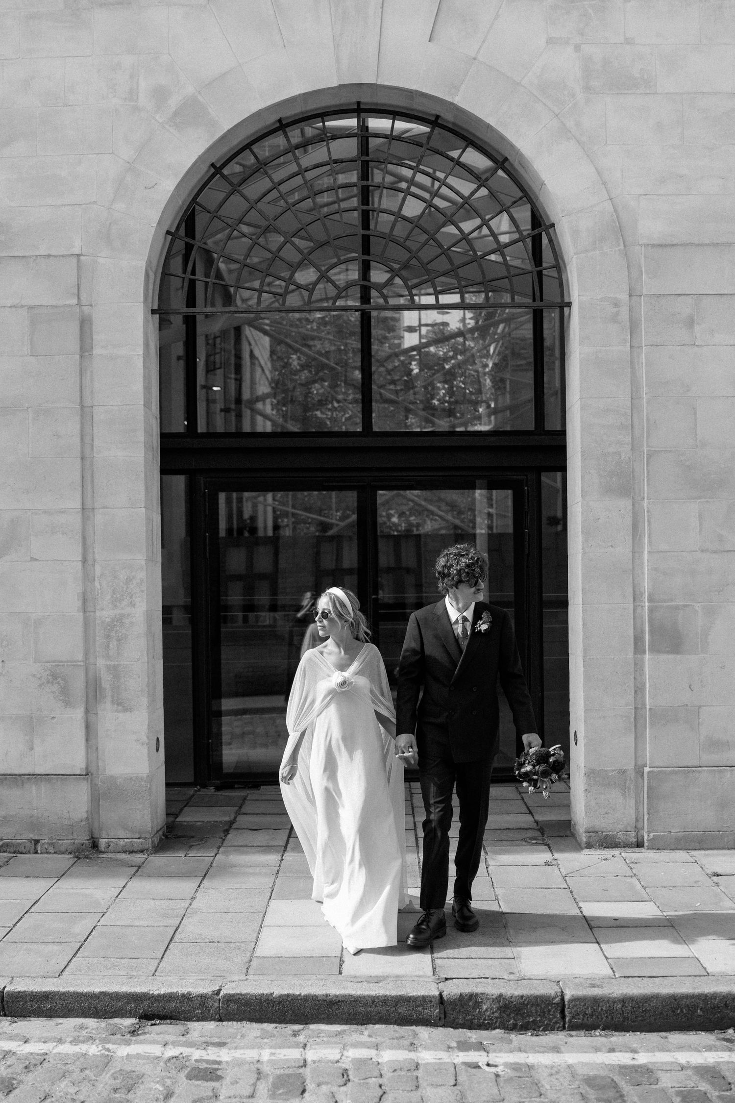 A black and white photo of newlyweds walking through an ornate arched entrance.