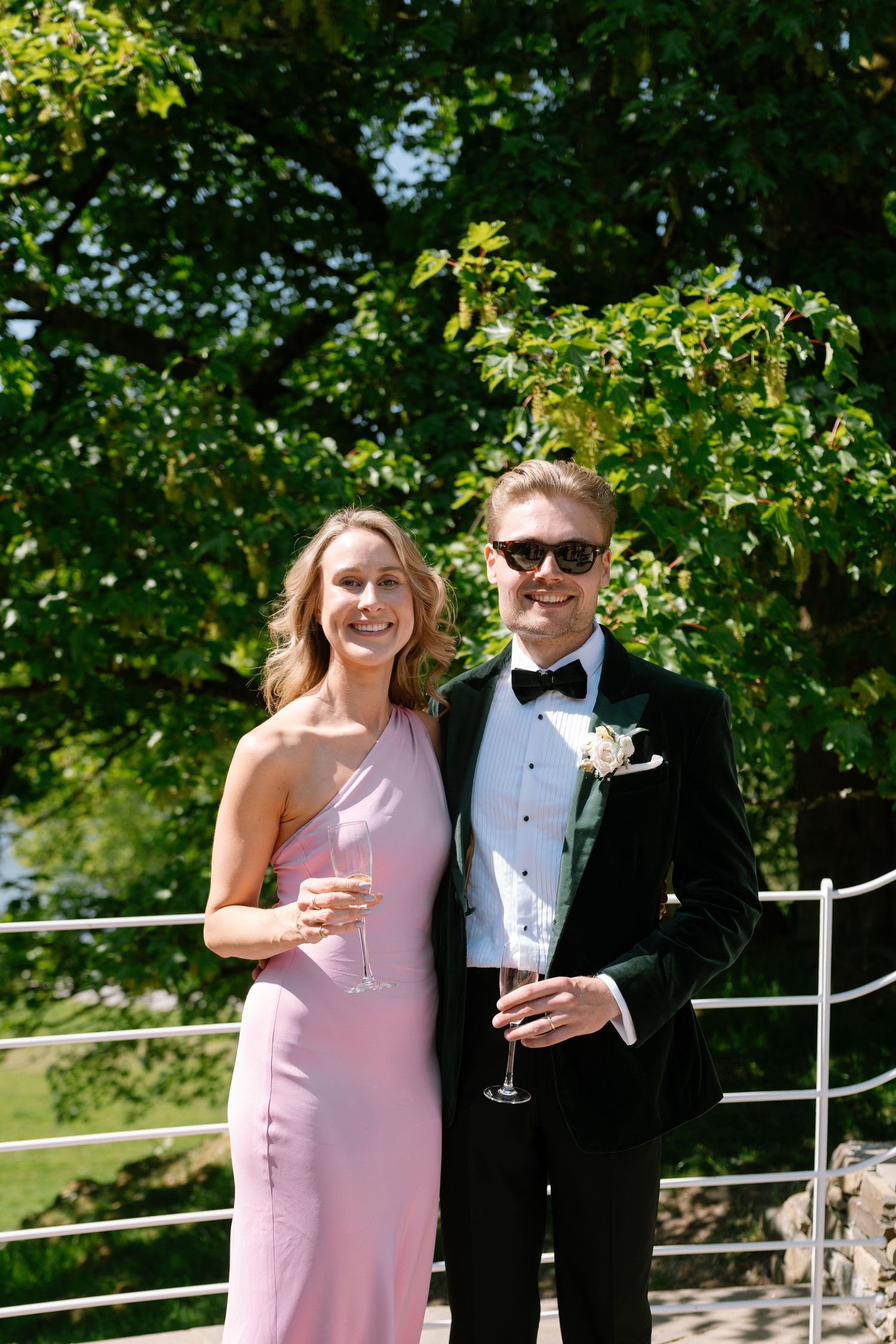 Couple in formal attire sharing champagne on a sunny terrace with white railings.