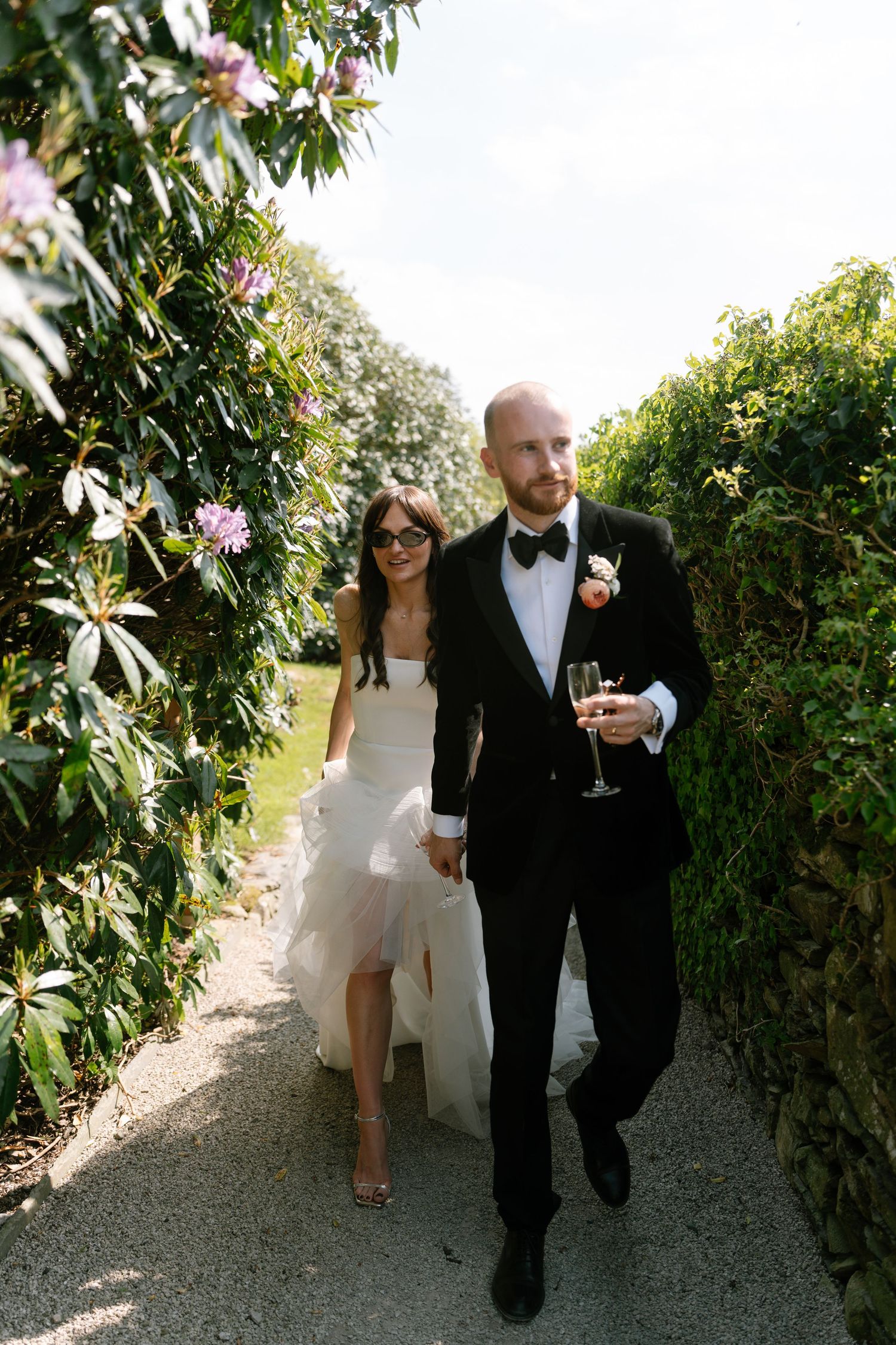 Newlyweds stroll along a garden path holding champagne glasses.