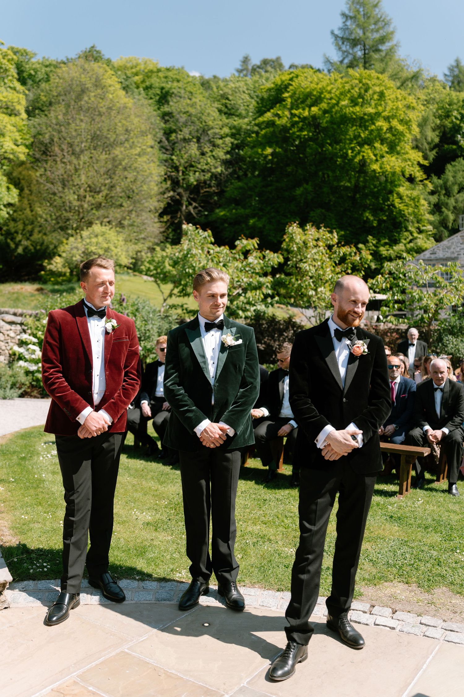 Groomsmen in tuxedos line up outdoors against a backdrop of green trees.