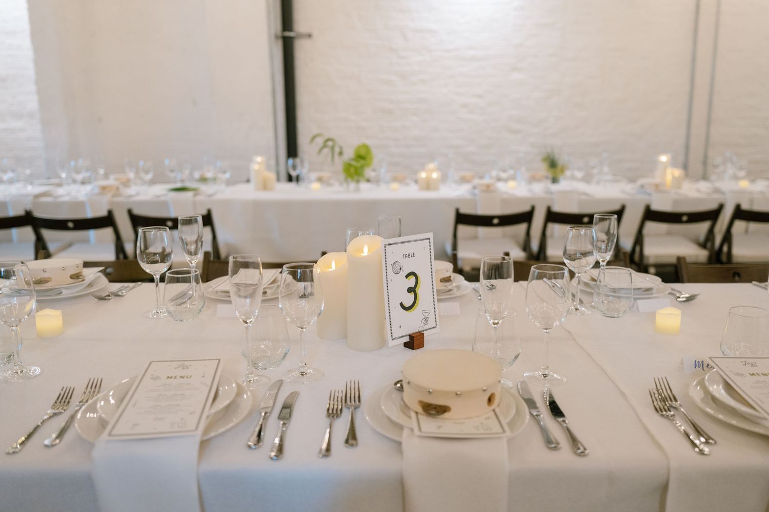 Elegant wedding reception tables set with white linens, candles and formal place settings in a bright venue.