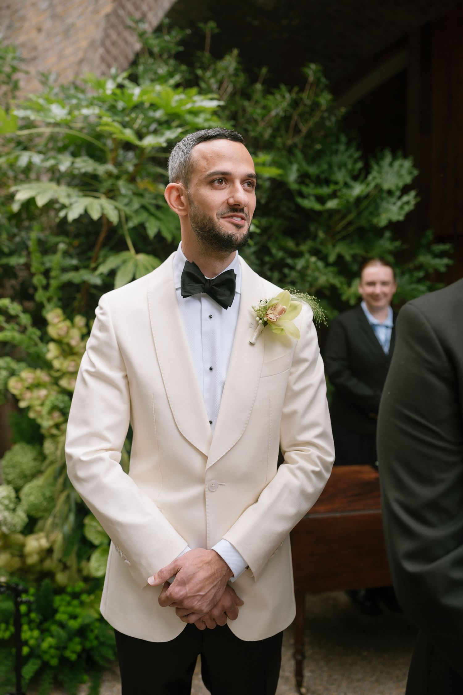 A groom in a white tuxedo jacket and black bow tie stands during a garden wedding ceremony.