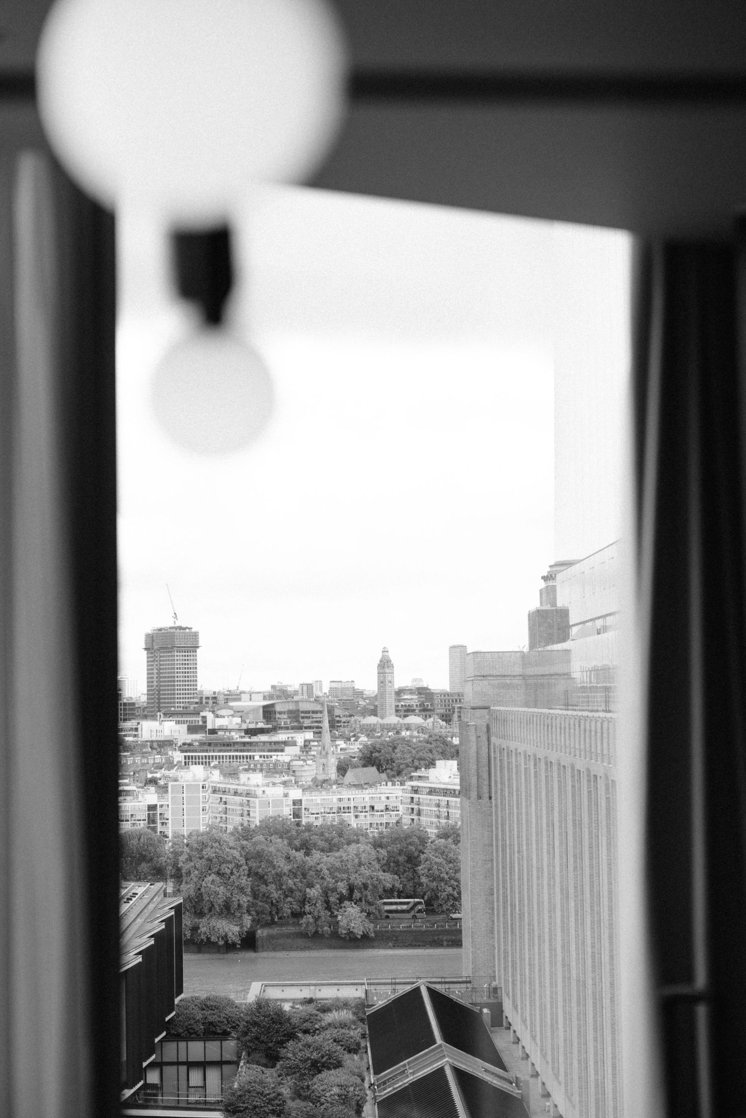 Black and white cityscape view through columns showing urban buildings and trees against an overcast sky.